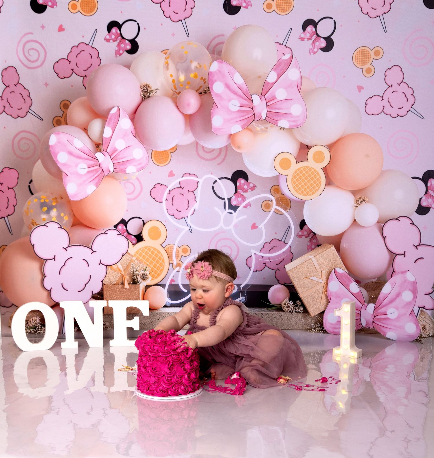 Baby in pink dress crawls and plays among pastel balloon arch with flowers in a cake smash studio photo session.
