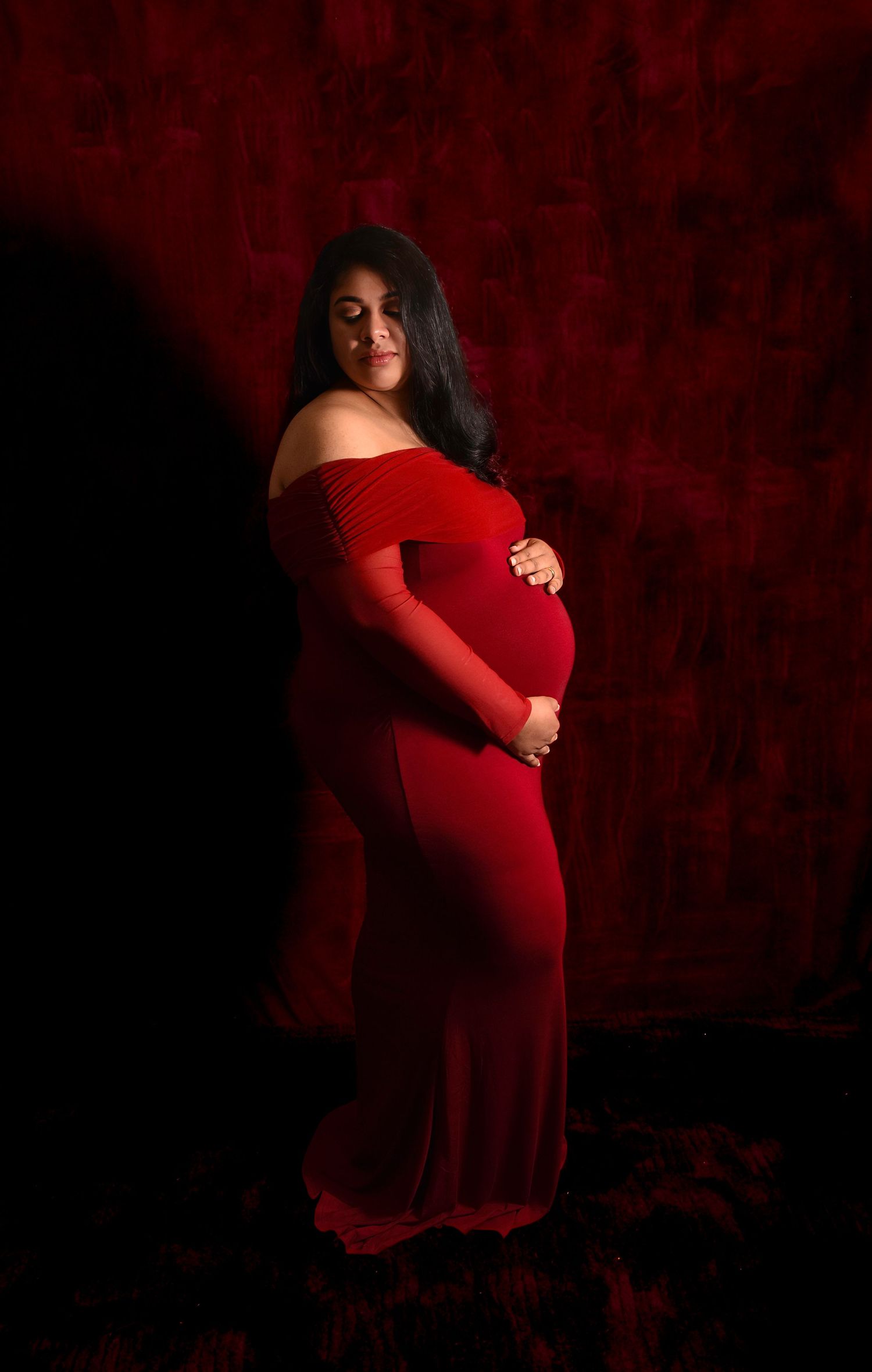 A maternity photoshoot featuring a woman in a flowing red dress against a dark dramatic background.