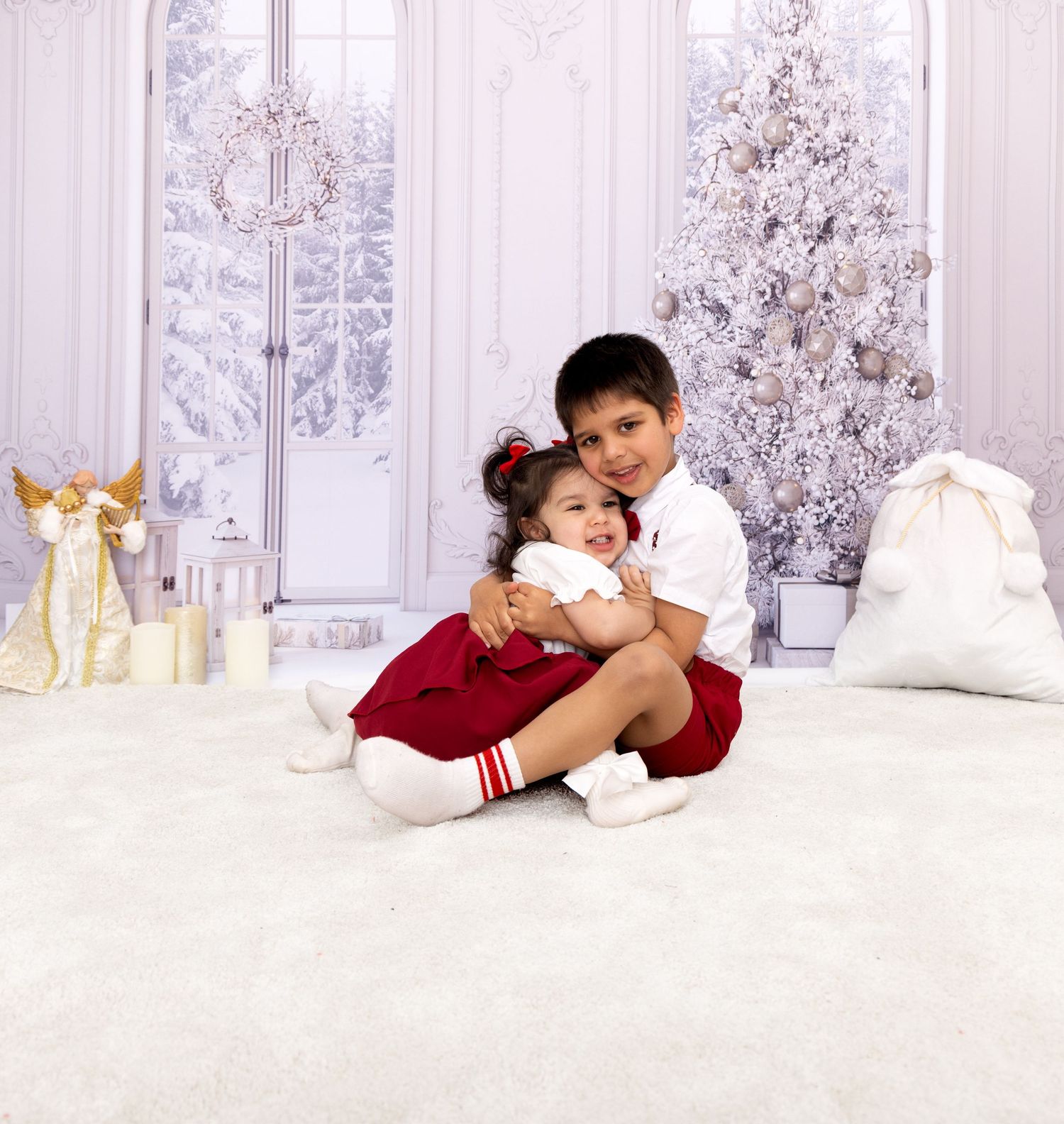Child in red velvet dress lying on white floor with Christmas decorations and snowy trees in background.