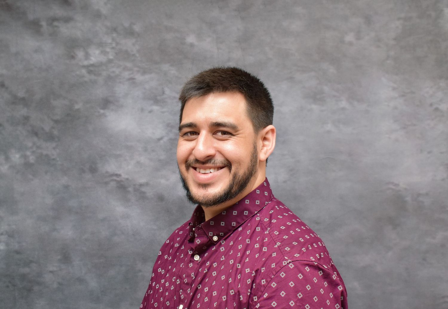 Professional headshot of person in burgundy polka dot shirt against textured wall.