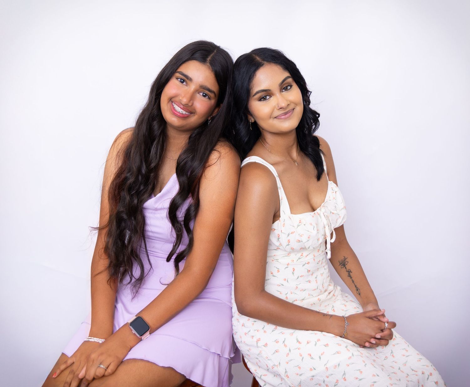 Two women in casual dresses pose together against a white background, one wearing lavender and one in white lace.