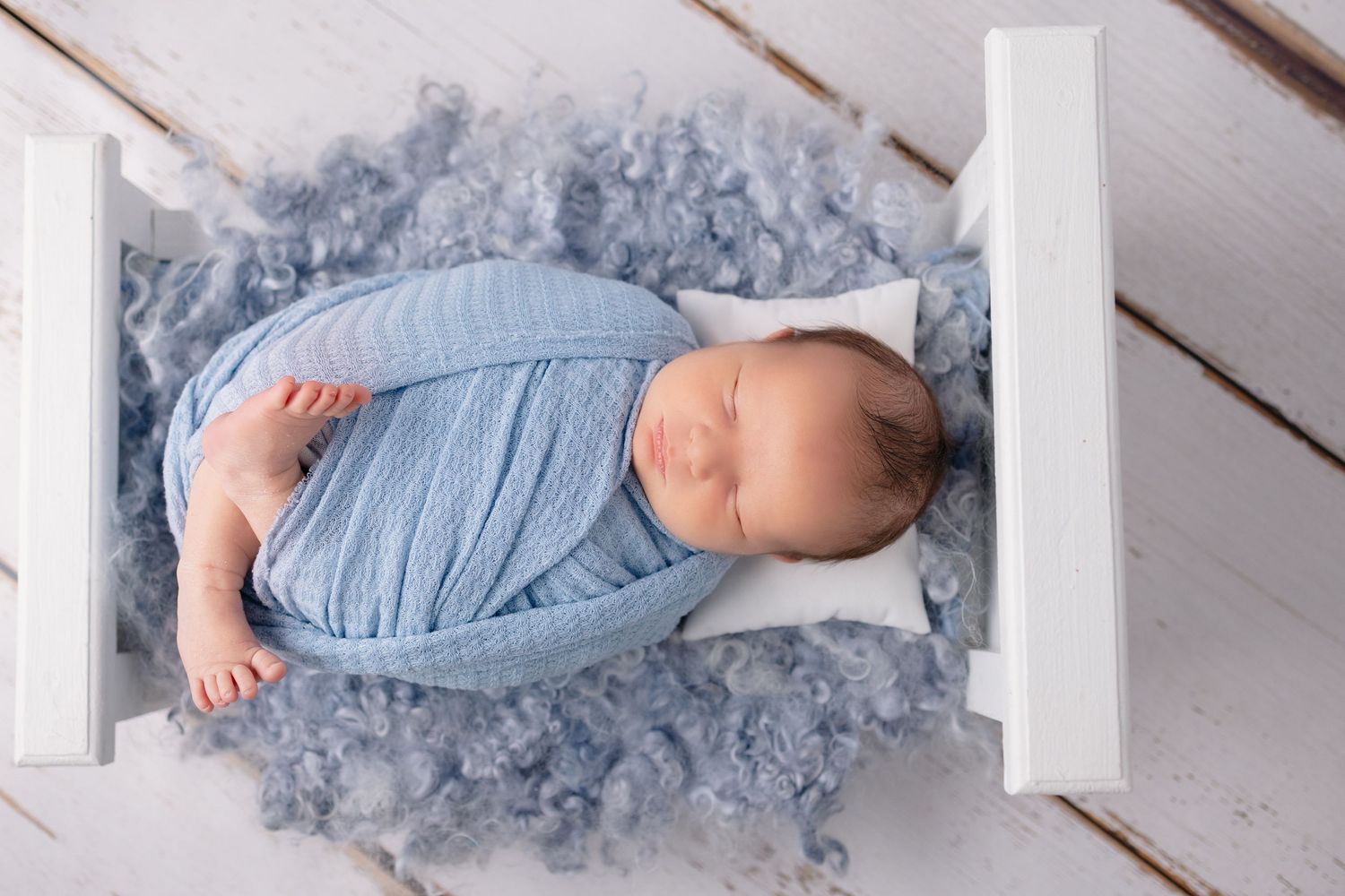 A baby in a blue dress lies on a white wooden floor while sleeping peacefully.