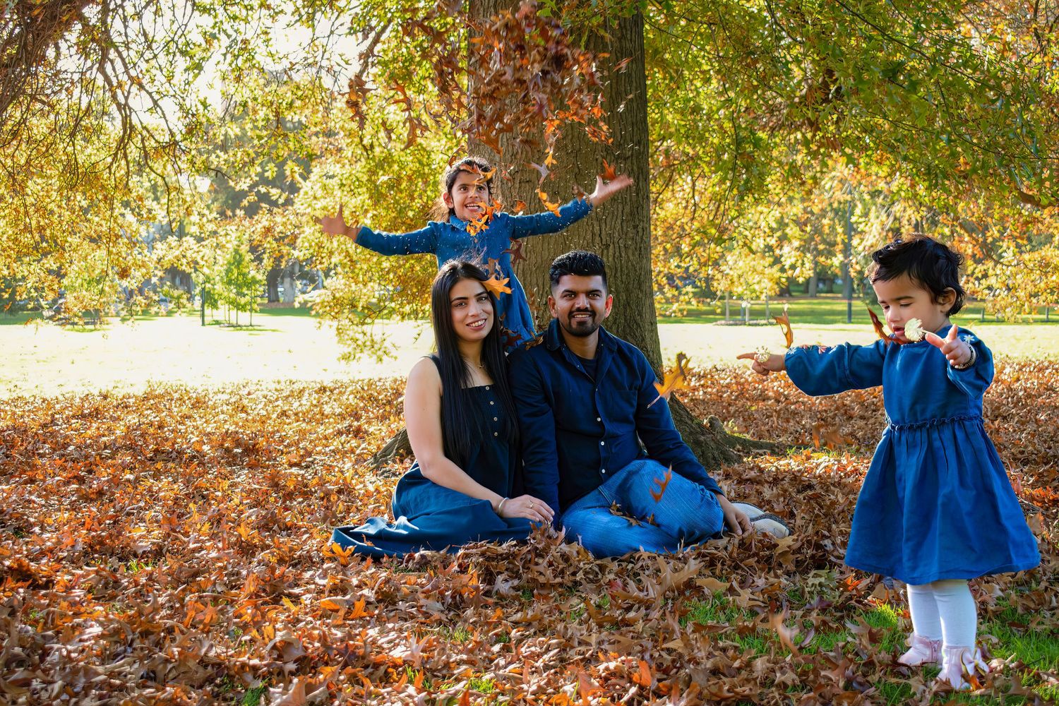 Family wearing coordinating blue outfits poses together during autumn photoshoot in park with fallen leaves.