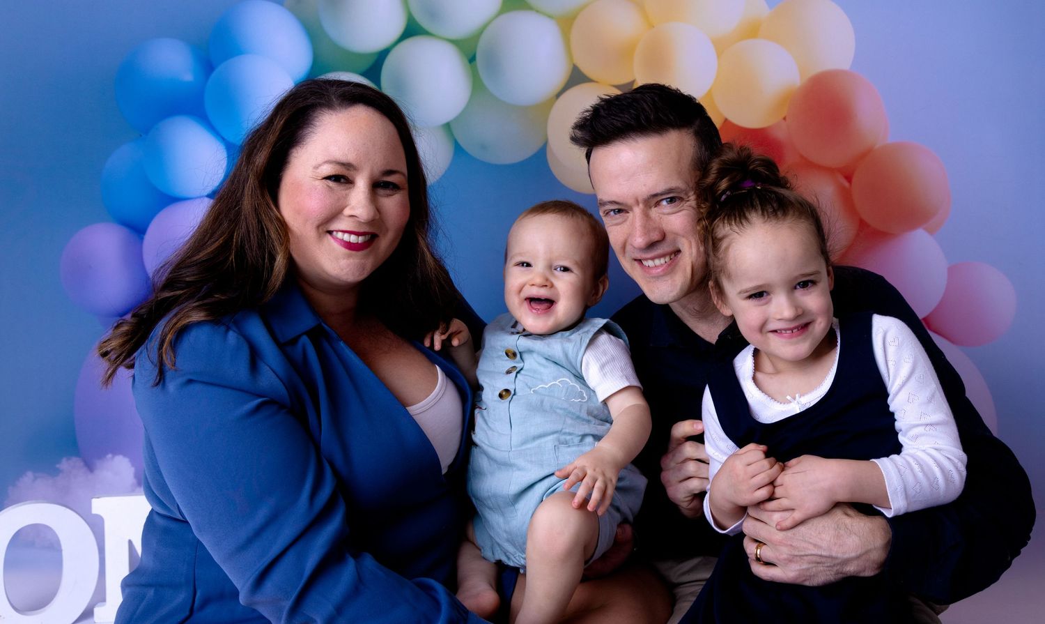 A family poses together against a colourful balloon arch backdrop during a mini photo session.
