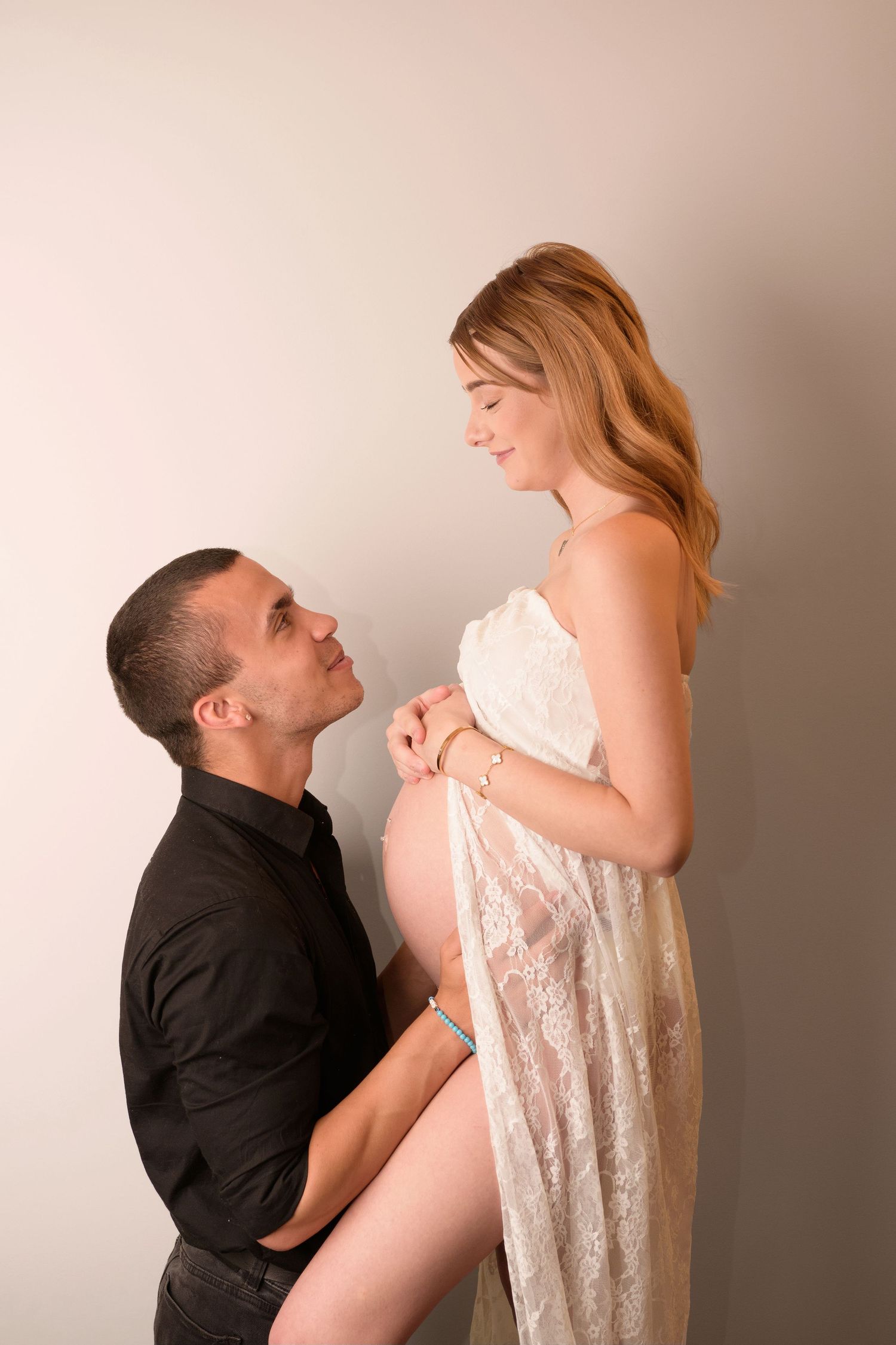 A couple in an intimate maternity photo shoot with the person wearing a black shirt kissing a white lace dress.