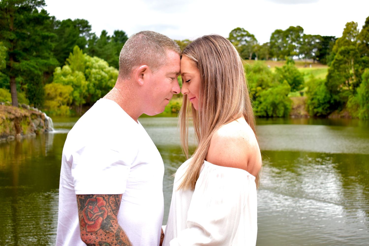 Romantic couple stands by a serene lake surrounded by lush greenery on a sunny afternoon.