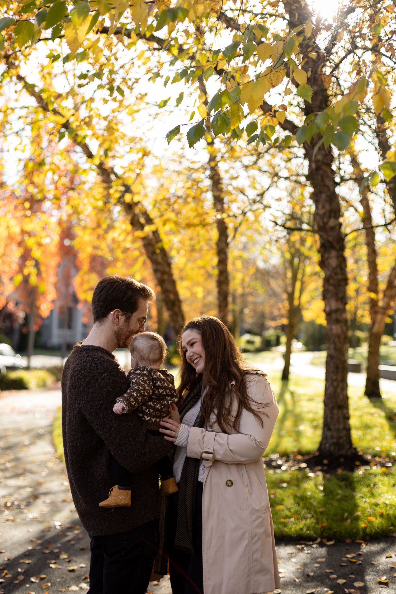 A family stands together on an autumn day with golden leaves and sunlight filtering through the trees.