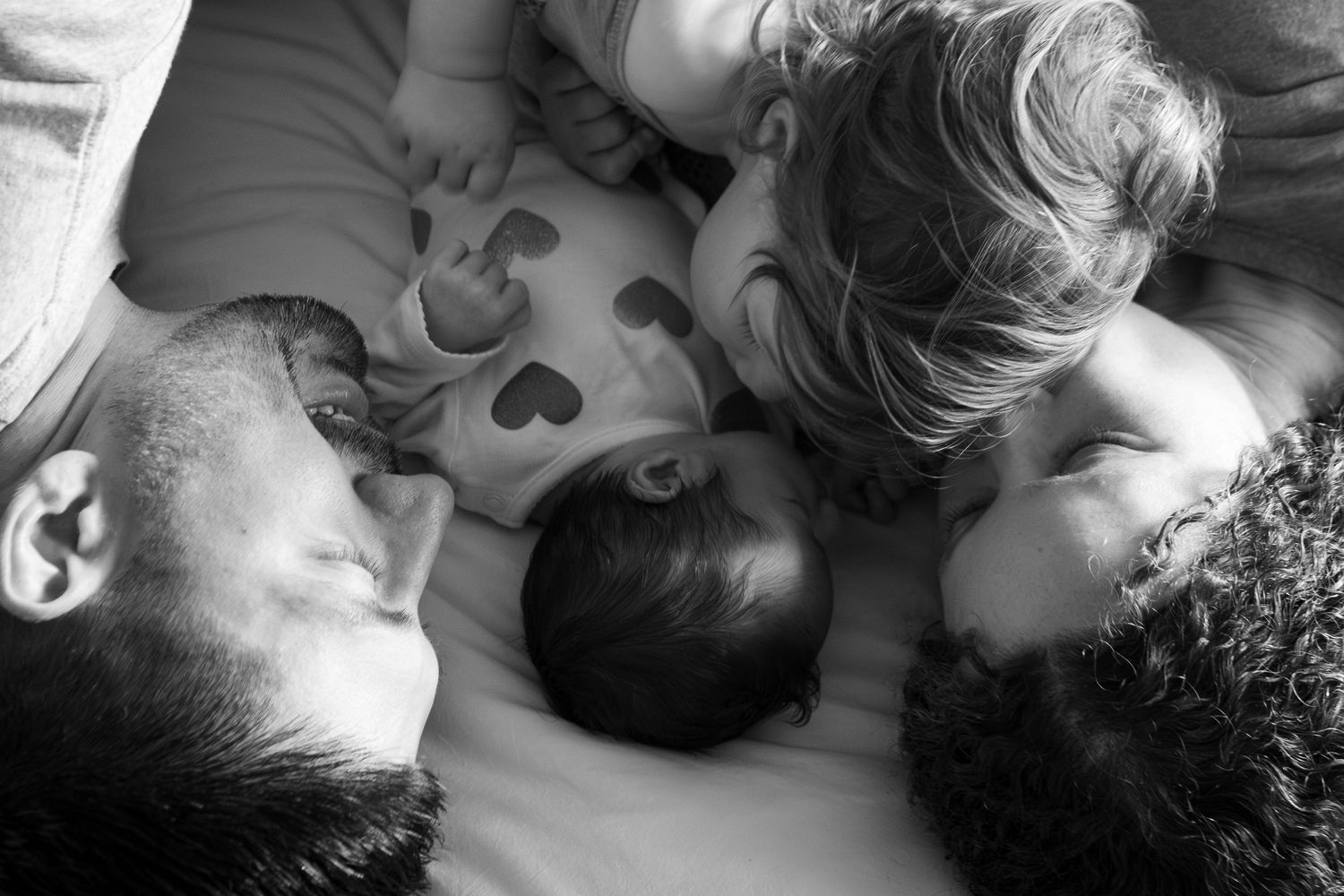 Black and white overhead view of family sharing intimate moment on heart-patterned bedding.