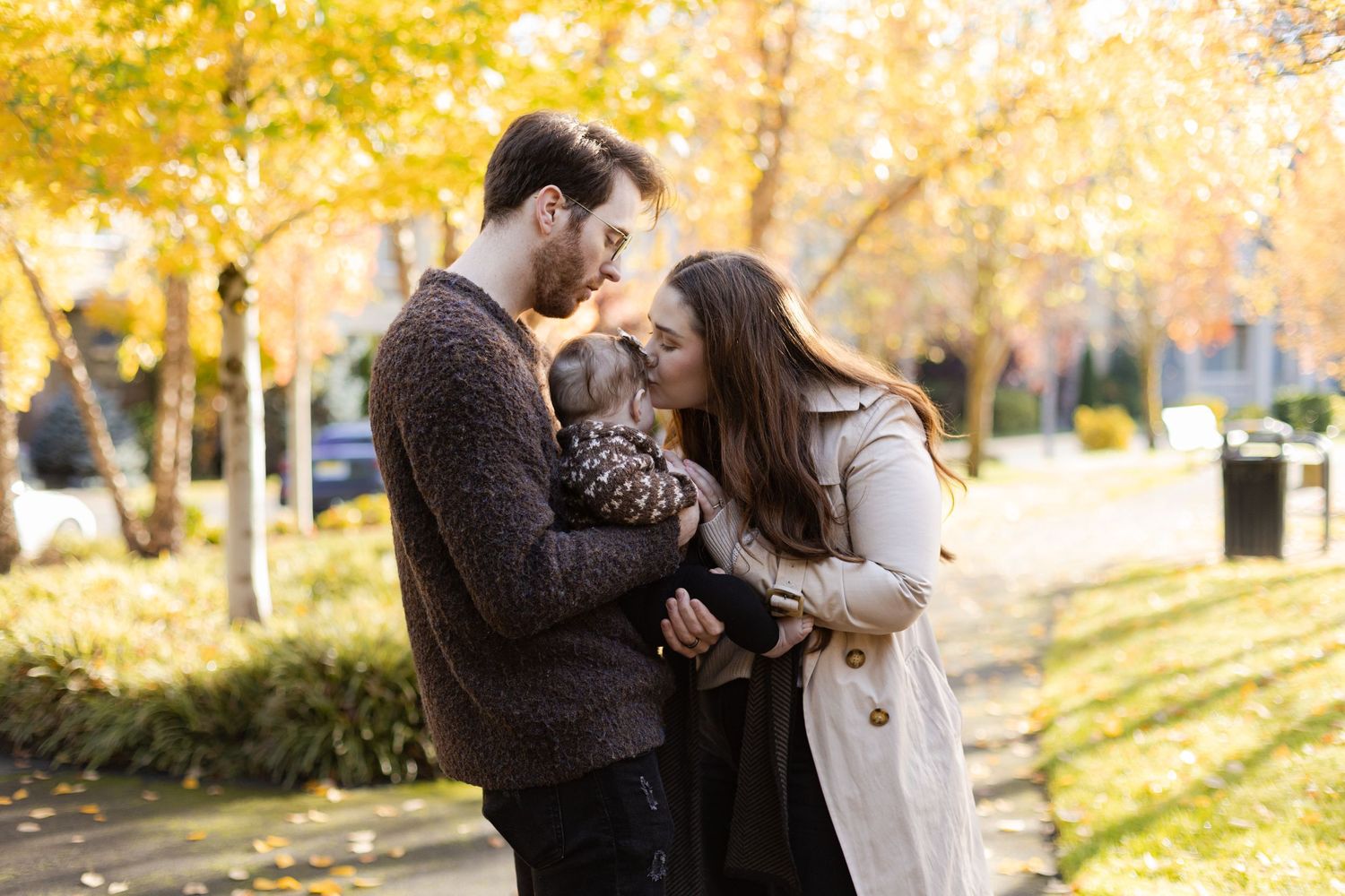 A couple holds a small baby together in a park with golden autumn foliage in the background on a sunny afternoon.