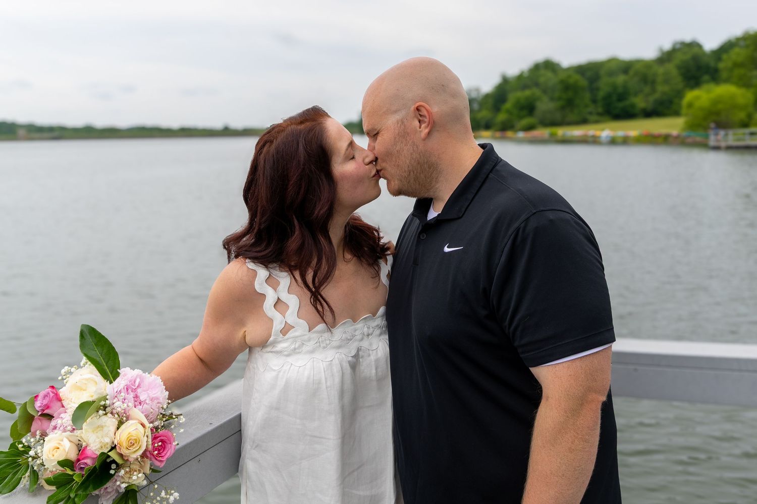 Couple sharing a romantic kiss on a waterfront deck, bride holding pink and white bouquet, groom in Nike polo.