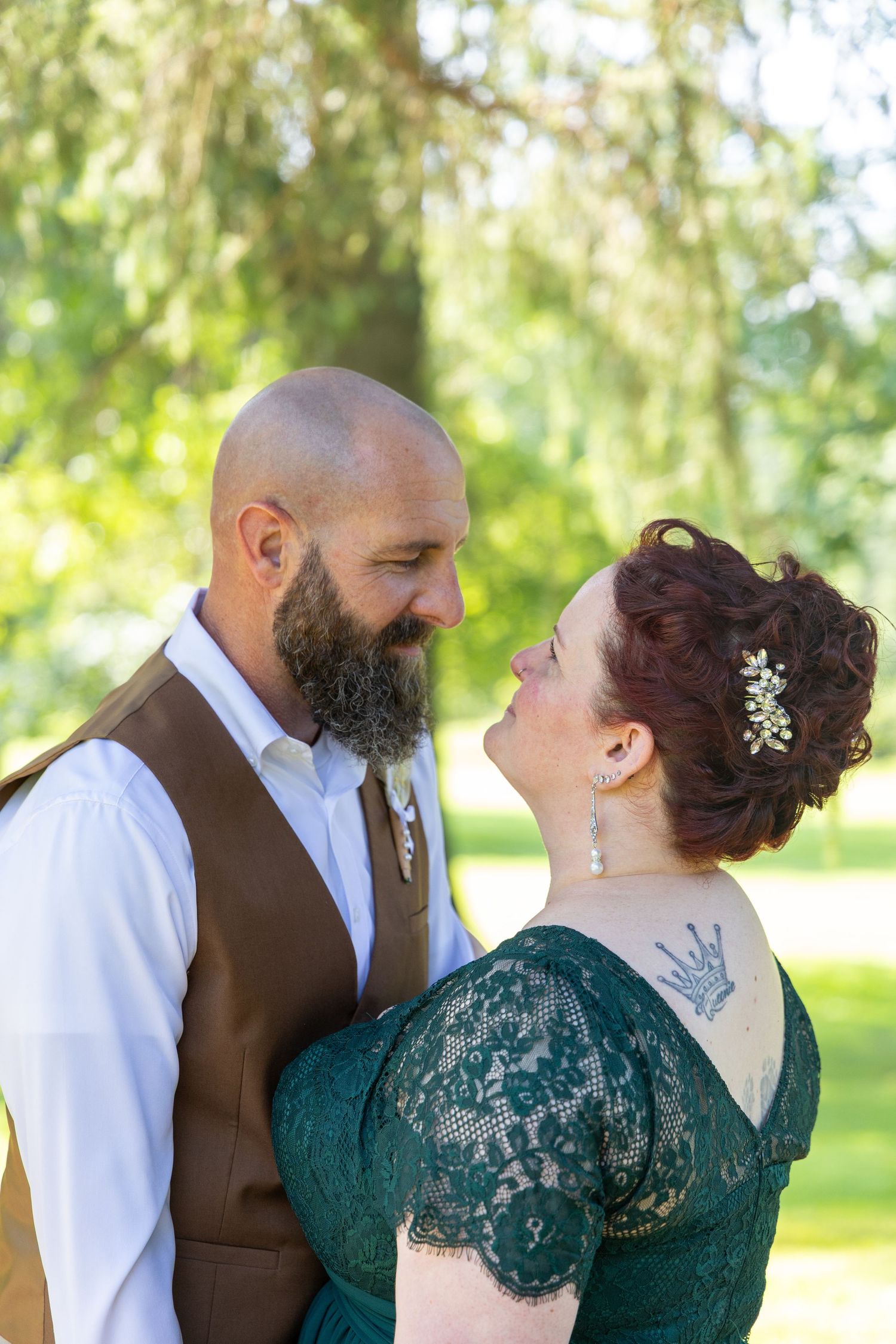 A couple shares a tender moment outdoors, with the woman wearing an emerald lace dress and the man in a brown vest.