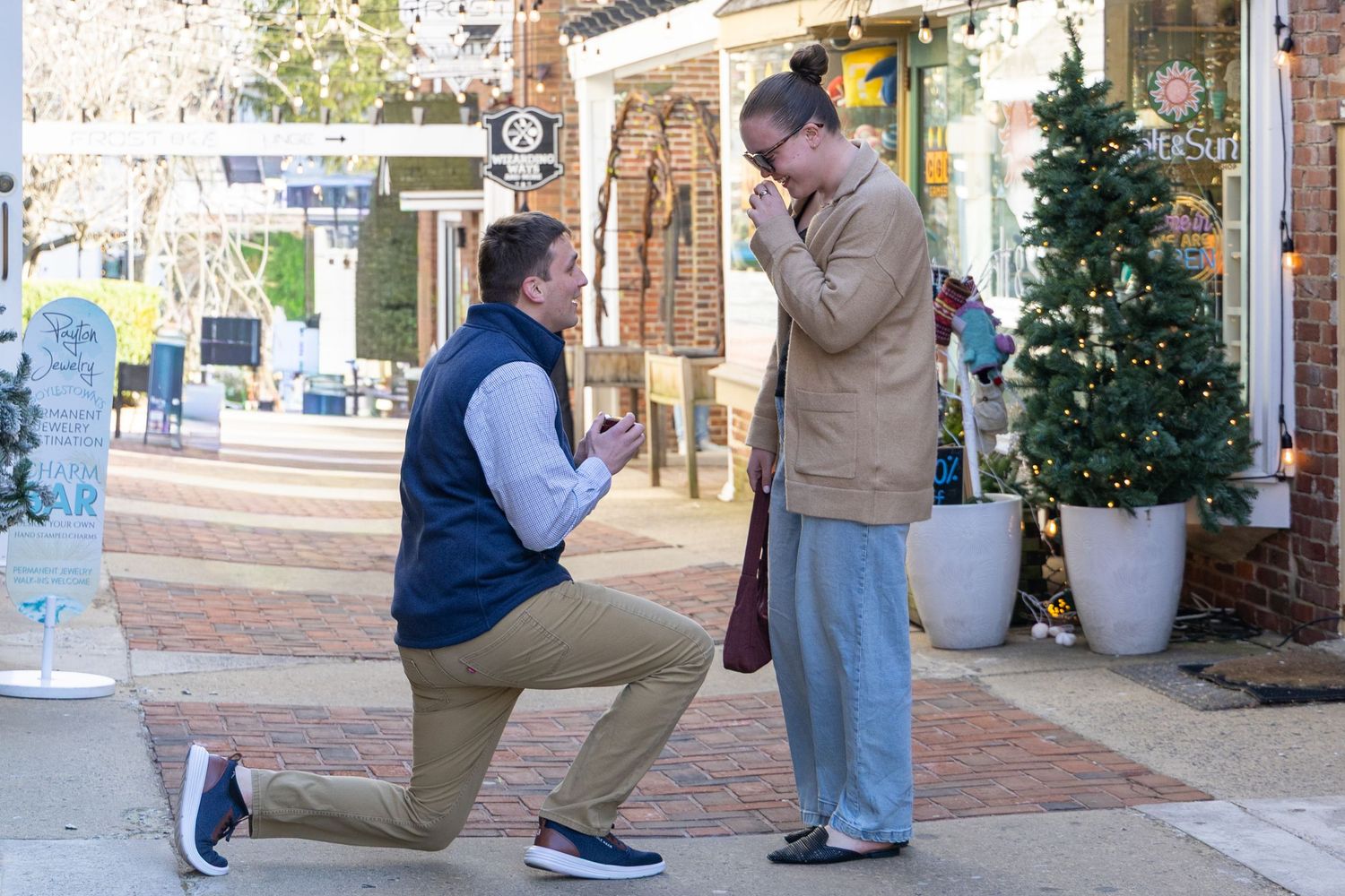 Man on one knee proposing to partner outdoors near Christmas tree in festive shopping district.