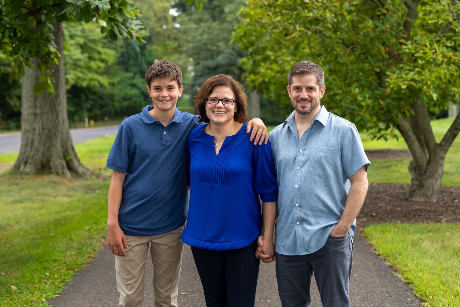 Family of three smiling on a tree-lined path outdoors, woman in blue top flanked by teenage boy and adult man.