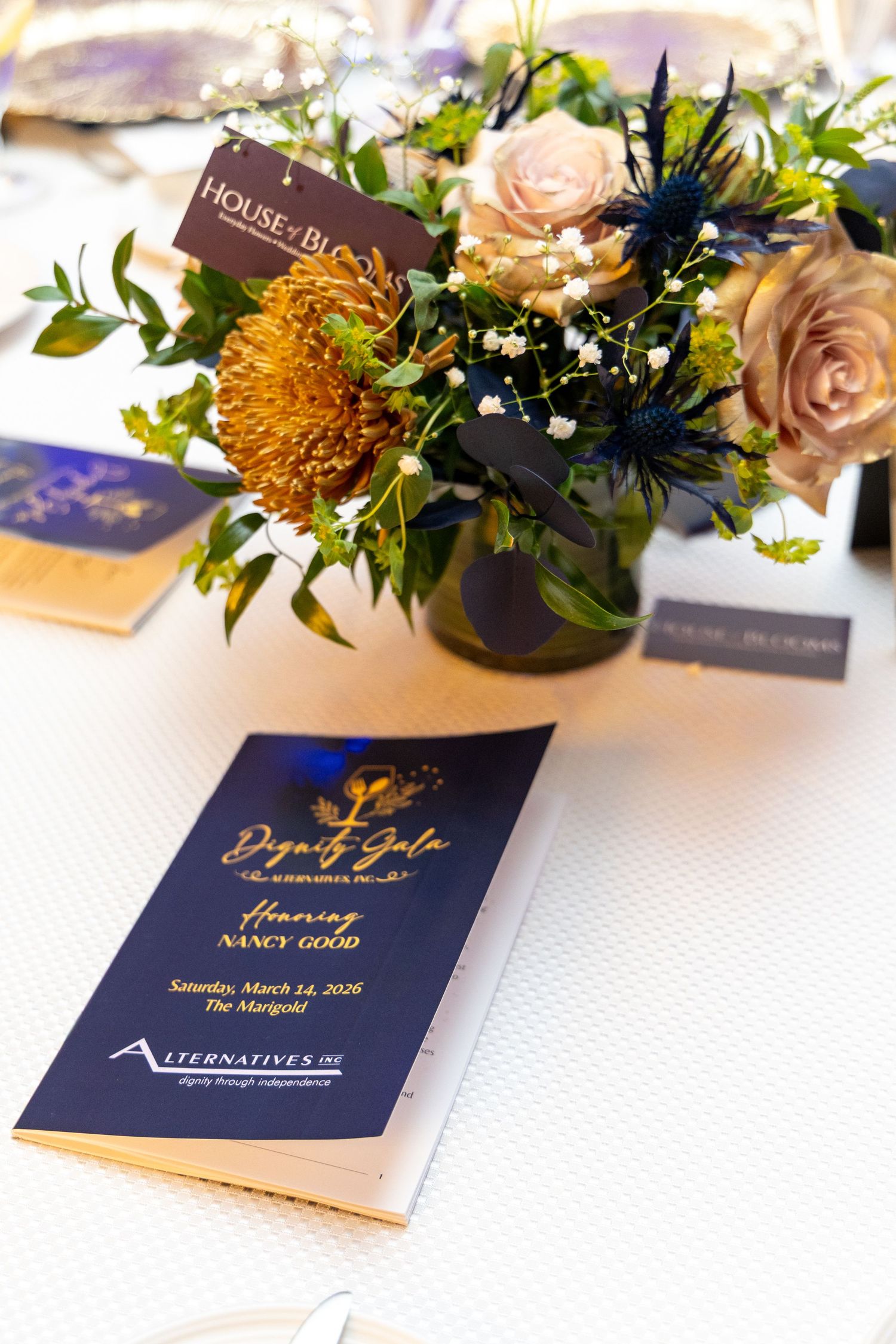Navy blue event program and floral centerpiece with peach roses and yellow chrysanthemums in glass vase on white tablecloth.