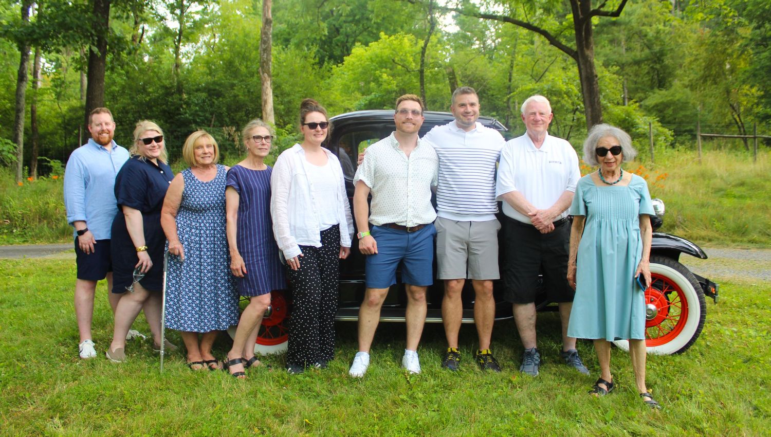 A casual group photo of people gathered outdoors near a vintage car on a summer day.