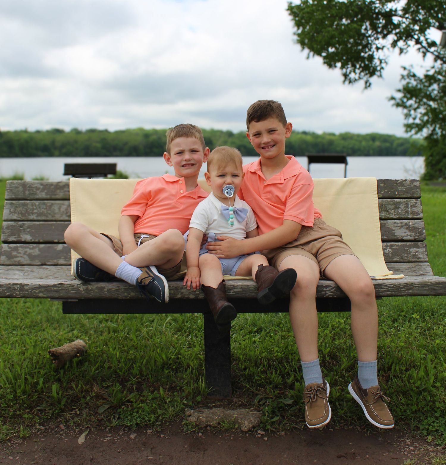 Three brothers in matching coral shirts sitting together on a park bench by a lake.
