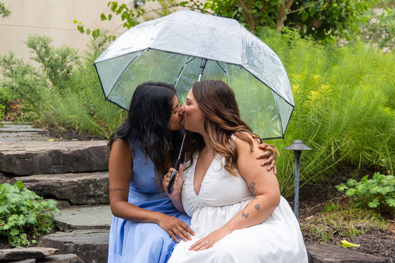 Two brides share a kiss under a clear umbrella in a lush garden, one in blue and one in white dress.