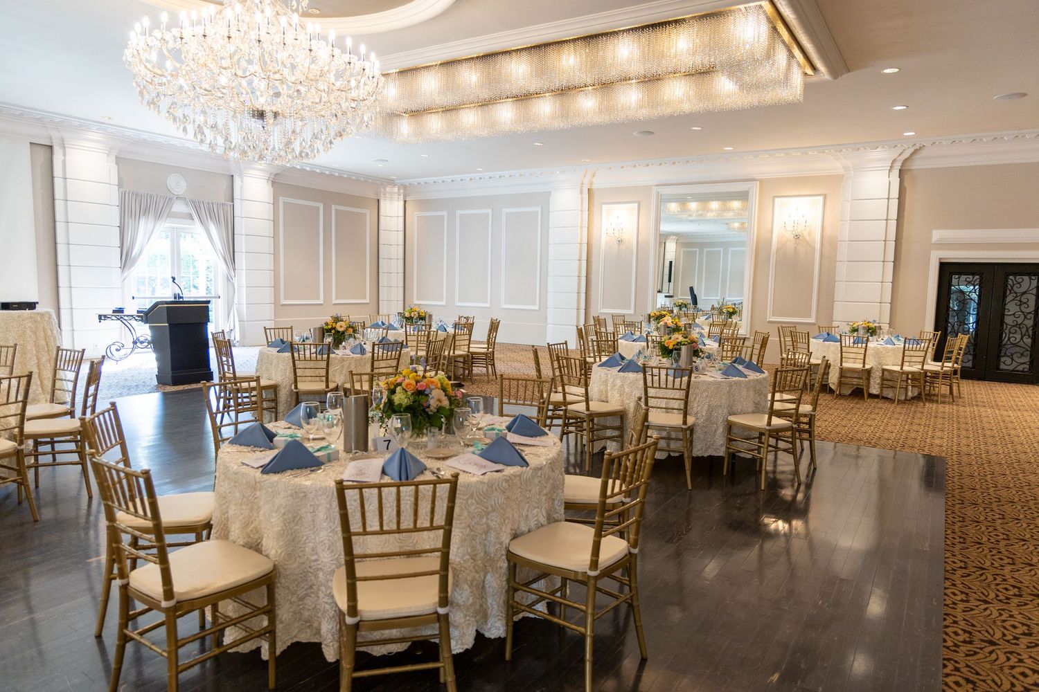 Elegant ballroom setup with round tables, gold chiavari chairs, crystal chandeliers and blue accents for a formal event.