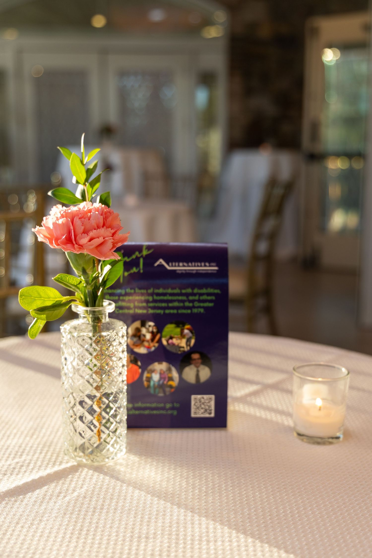 Small pink flower in crystal vase with candlelight and blue card on white tablecloth at elegant event setting.