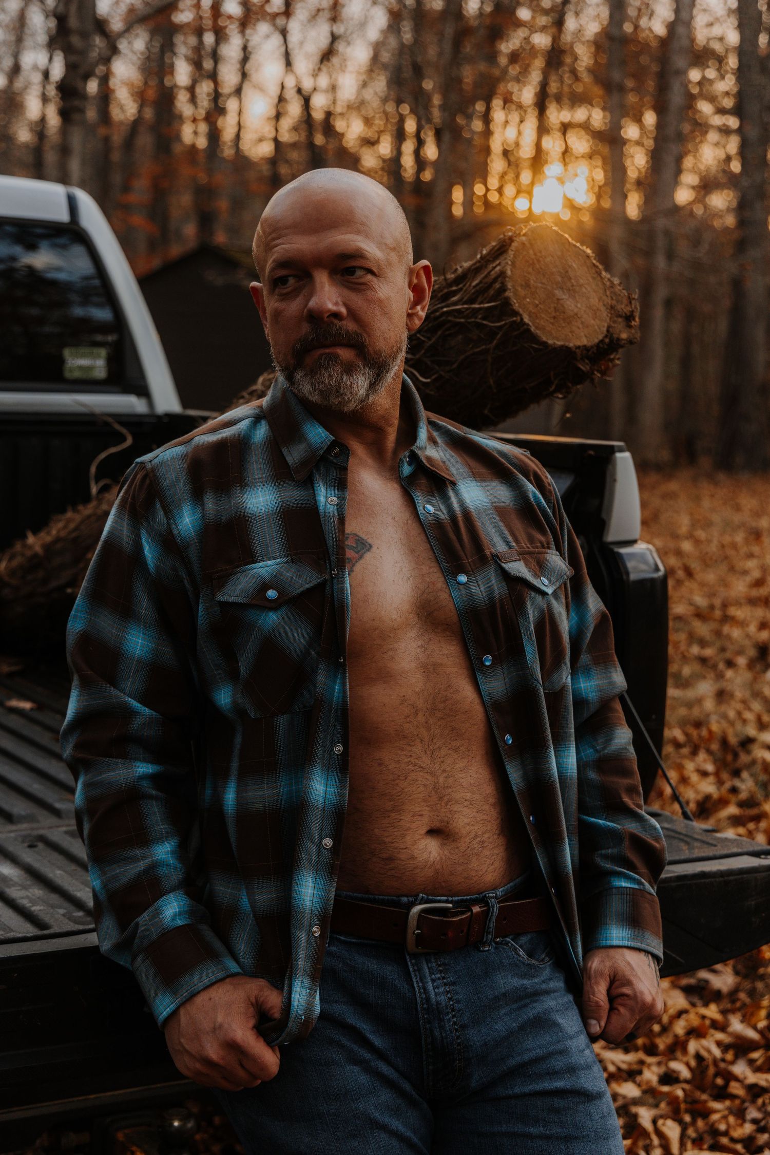 A rugged figure in a blue plaid flannel shirt poses against an autumn forest backdrop during golden hour.