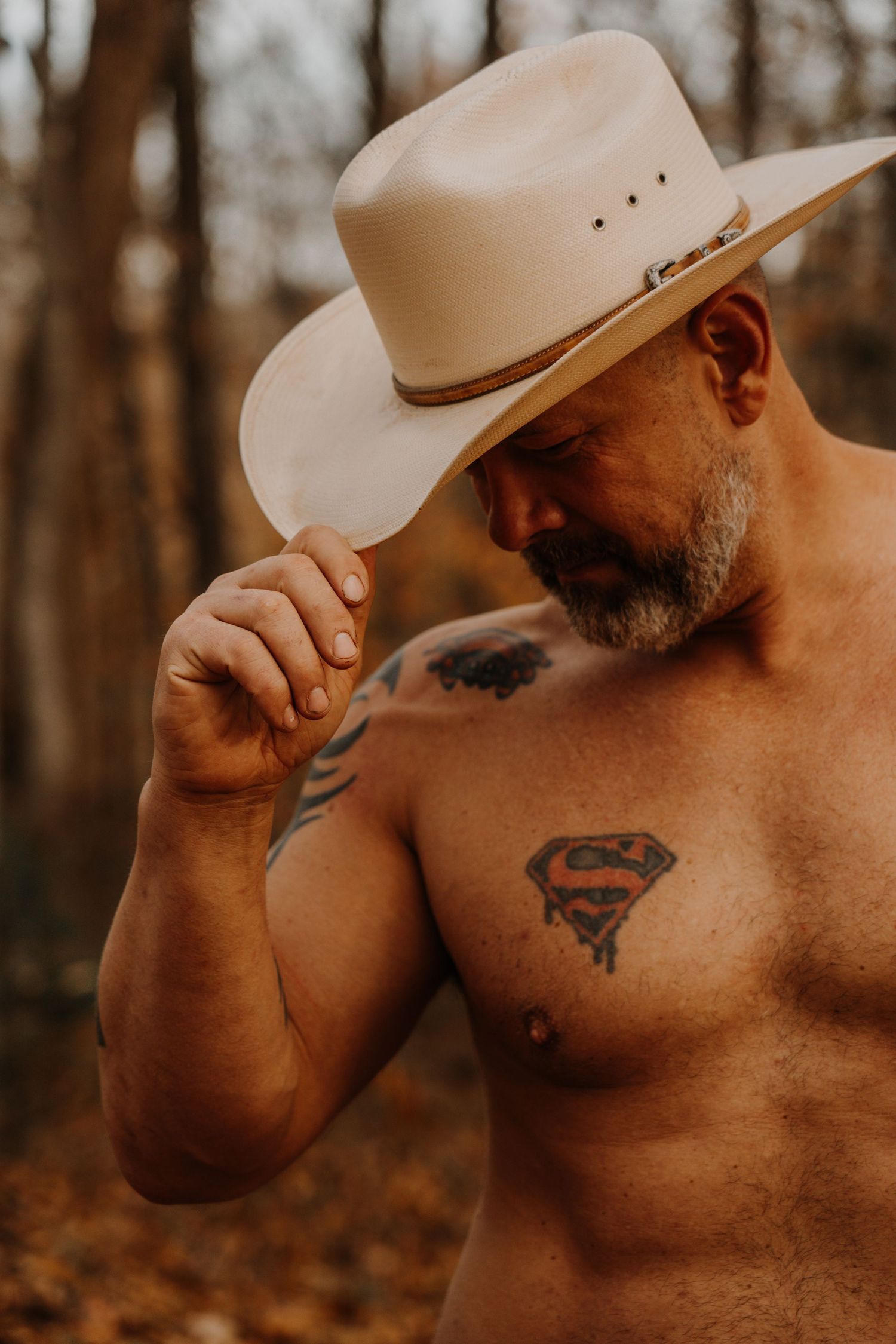 Shirtless man in cowboy hat with tattoos poses outdoors in autumn forest setting.