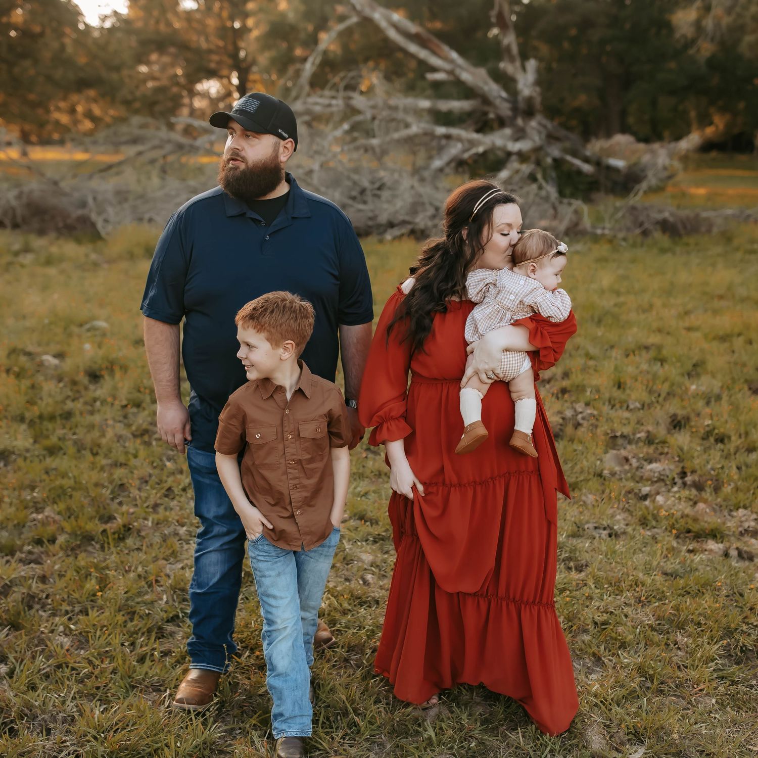 Family in rustic autumn setting with woman in flowing red dress posing together in grassy field at sunset.