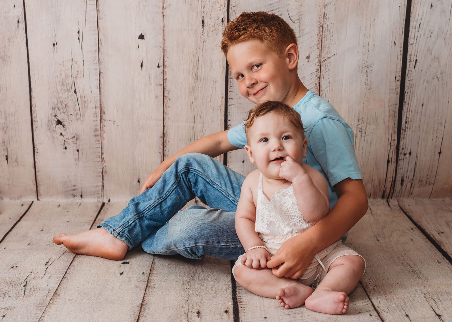 Two children in casual clothing pose together against a rustic white wooden wall backdrop.