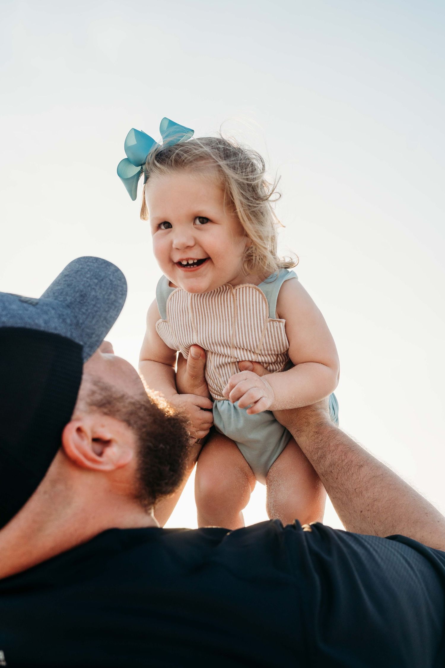 A playful moment captured as a child wearing a blue bow and striped top is lifted up against a bright sky.