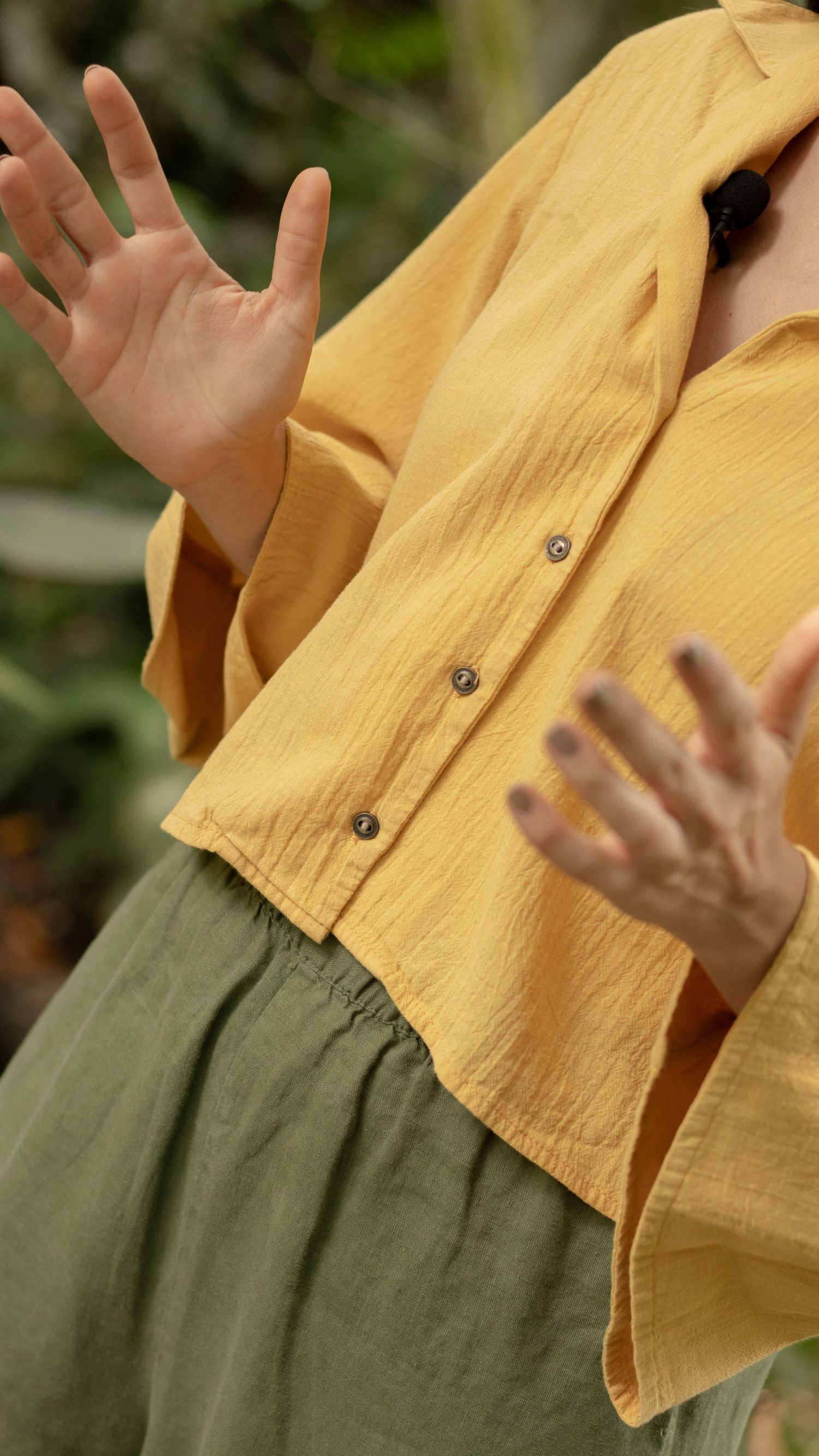 Person wearing yellow button-up shirt and olive skirt with gesturing hands.