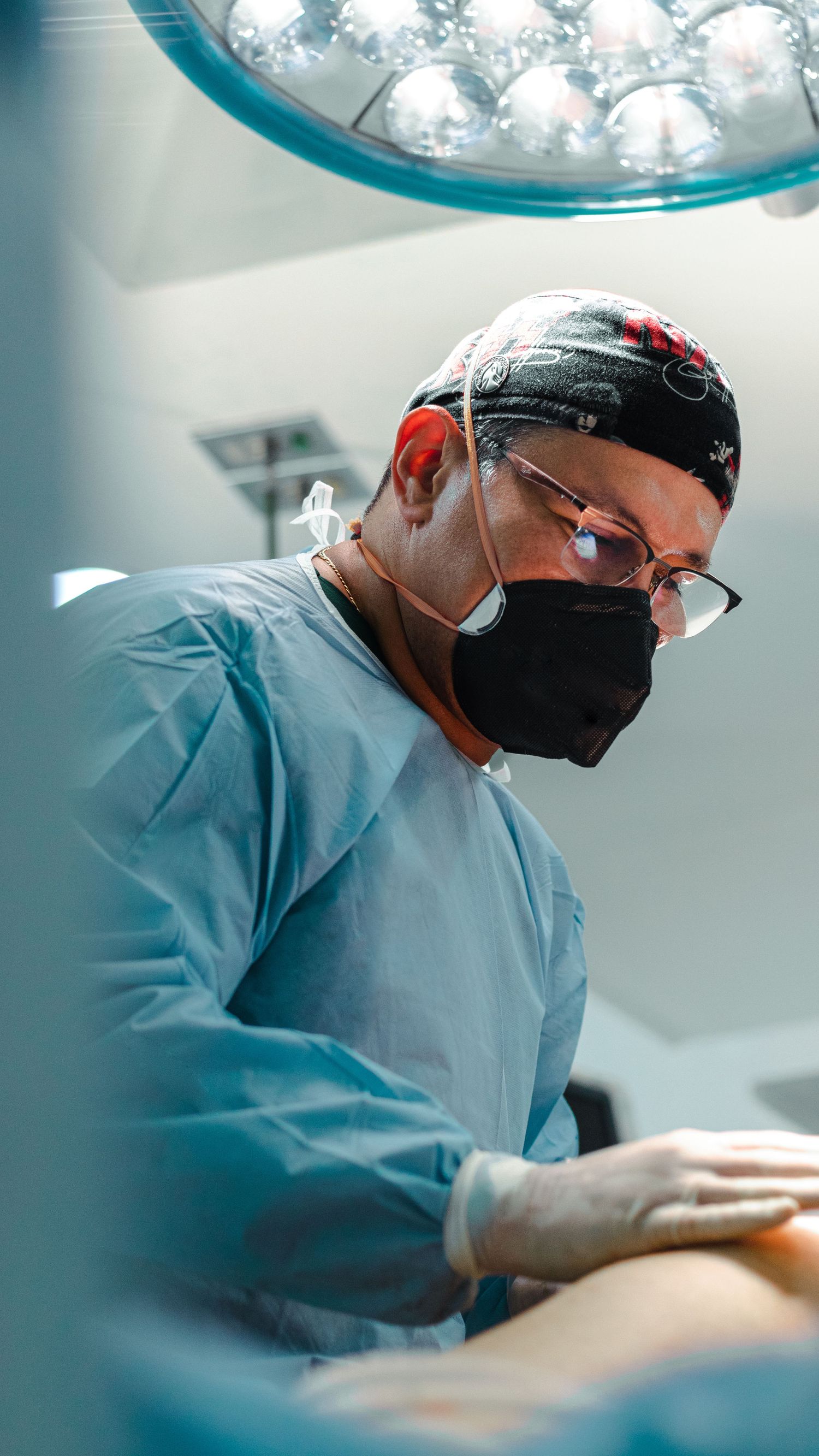 Surgeon in protective gear concentrating during medical procedure under surgical lighting.
