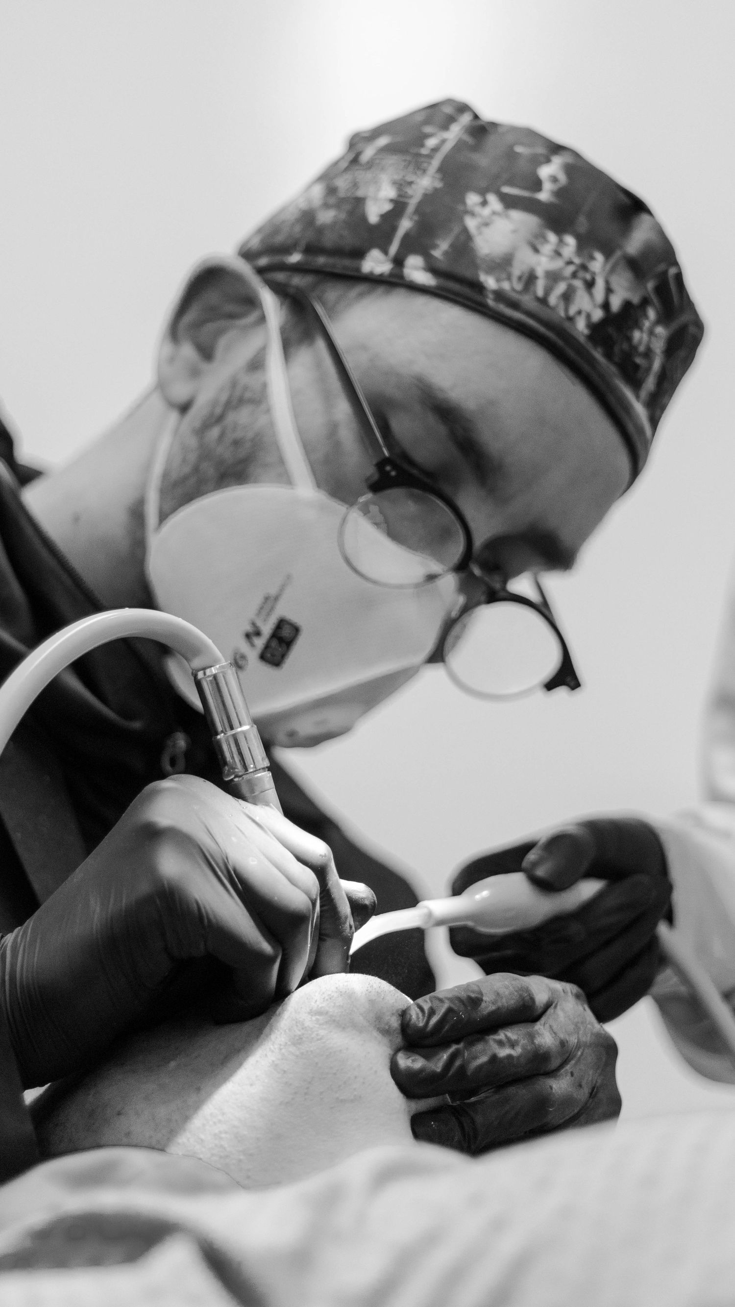 Medical professional wearing protective gear and glasses performs a dental procedure, viewed in black and white.
