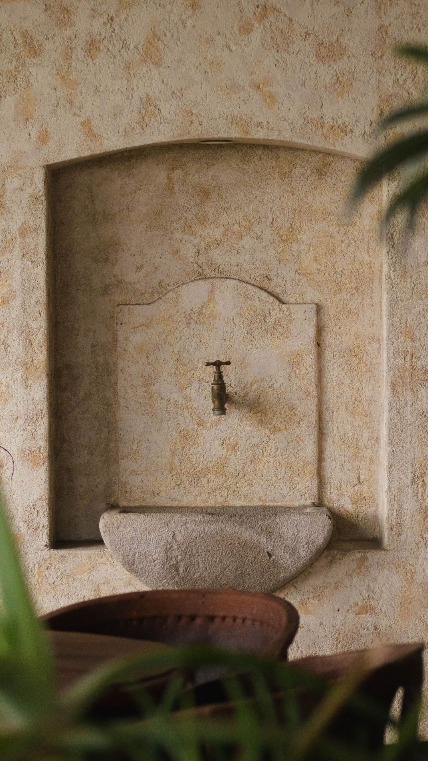 Rustic stone water fountain with basin mounted in beige stucco wall alcove.