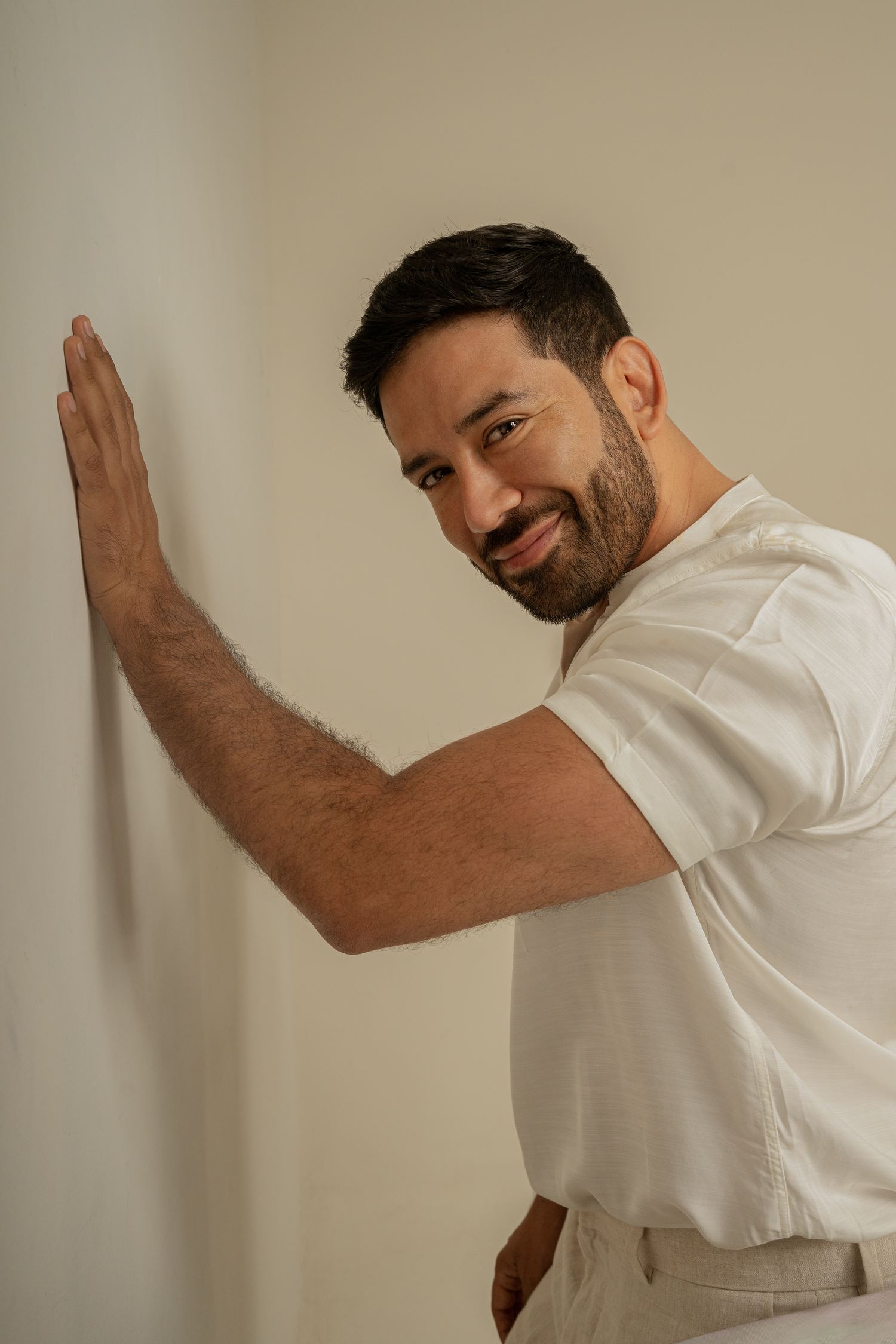 Man in white t-shirt smiling while leaning against wall in casual pose.