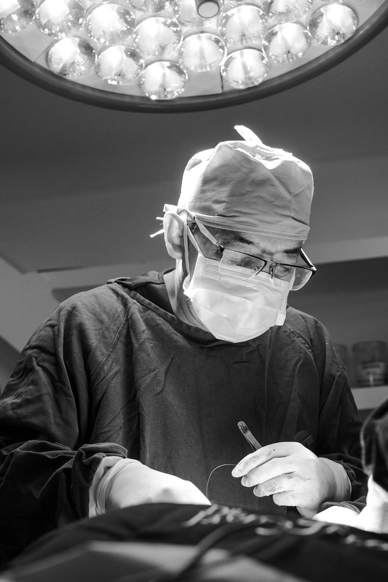 Surgeon operating under bright surgical lights in black and white photograph.