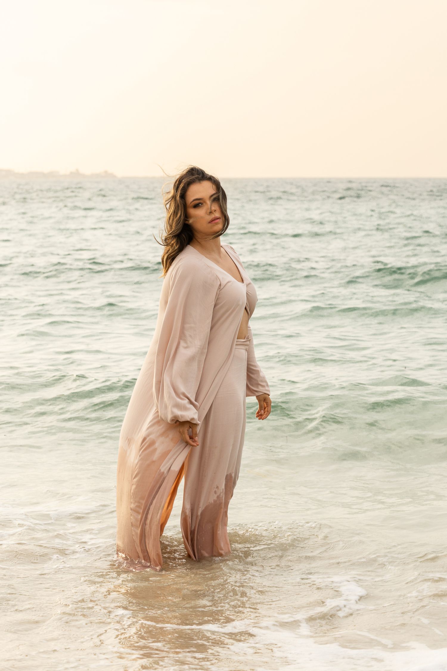 Ethereal beachwear shoot with flowing nude dress in gentle ocean waves at sunset.