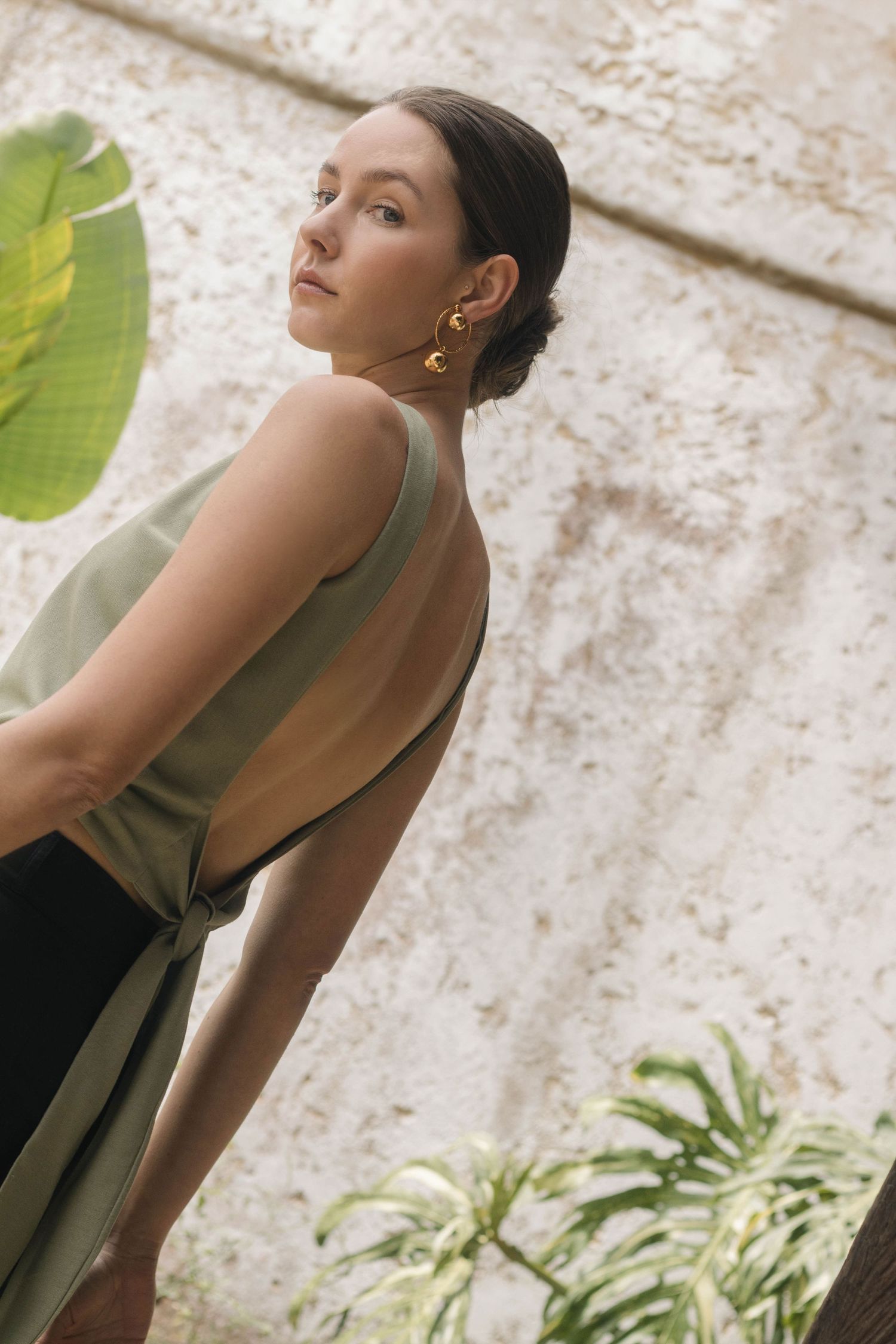 Fashion model in green top poses against textured wall with dramatic shadows and tropical plants.