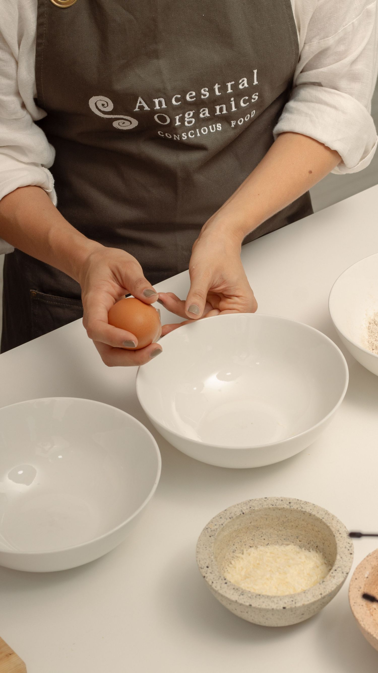 Hands wearing brown apron cracking an egg into white ceramic bowls on kitchen counter.