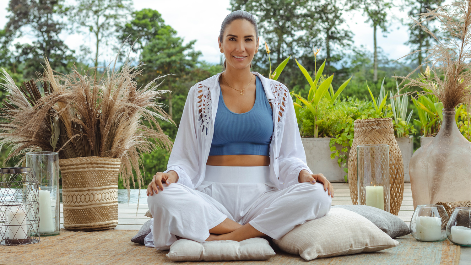 Person in white meditation outfit sitting on cushions in tropical outdoor setting.