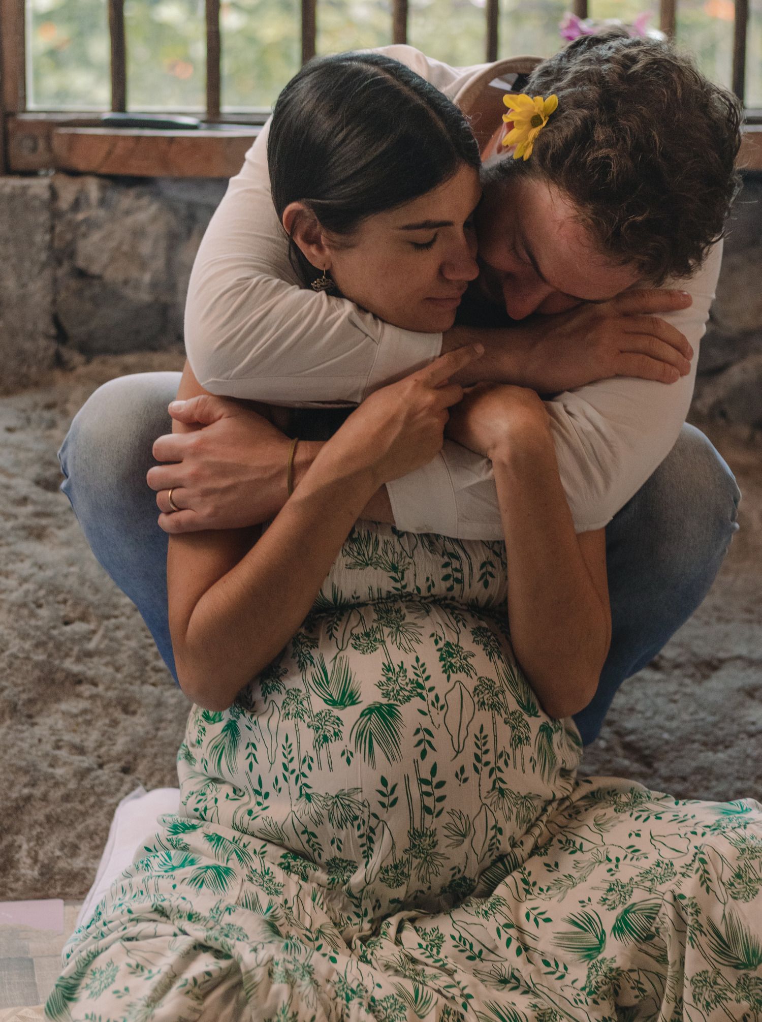 Two people share a tender embrace while sitting together, one wearing a floral patterned dress.