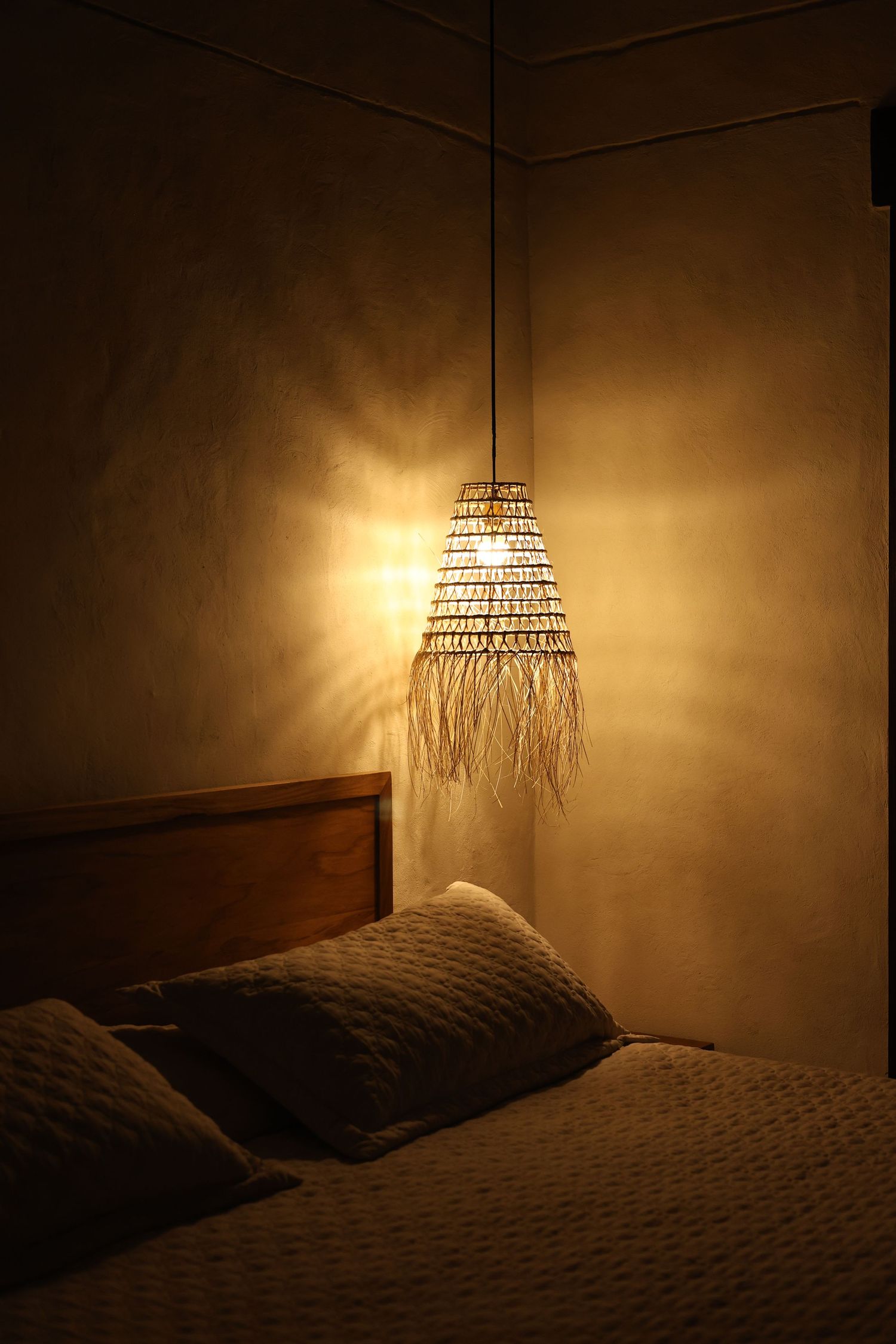 Cozy bedroom with warm glowing wicker pendant lamp casting golden light on textured wall above wooden headboard.