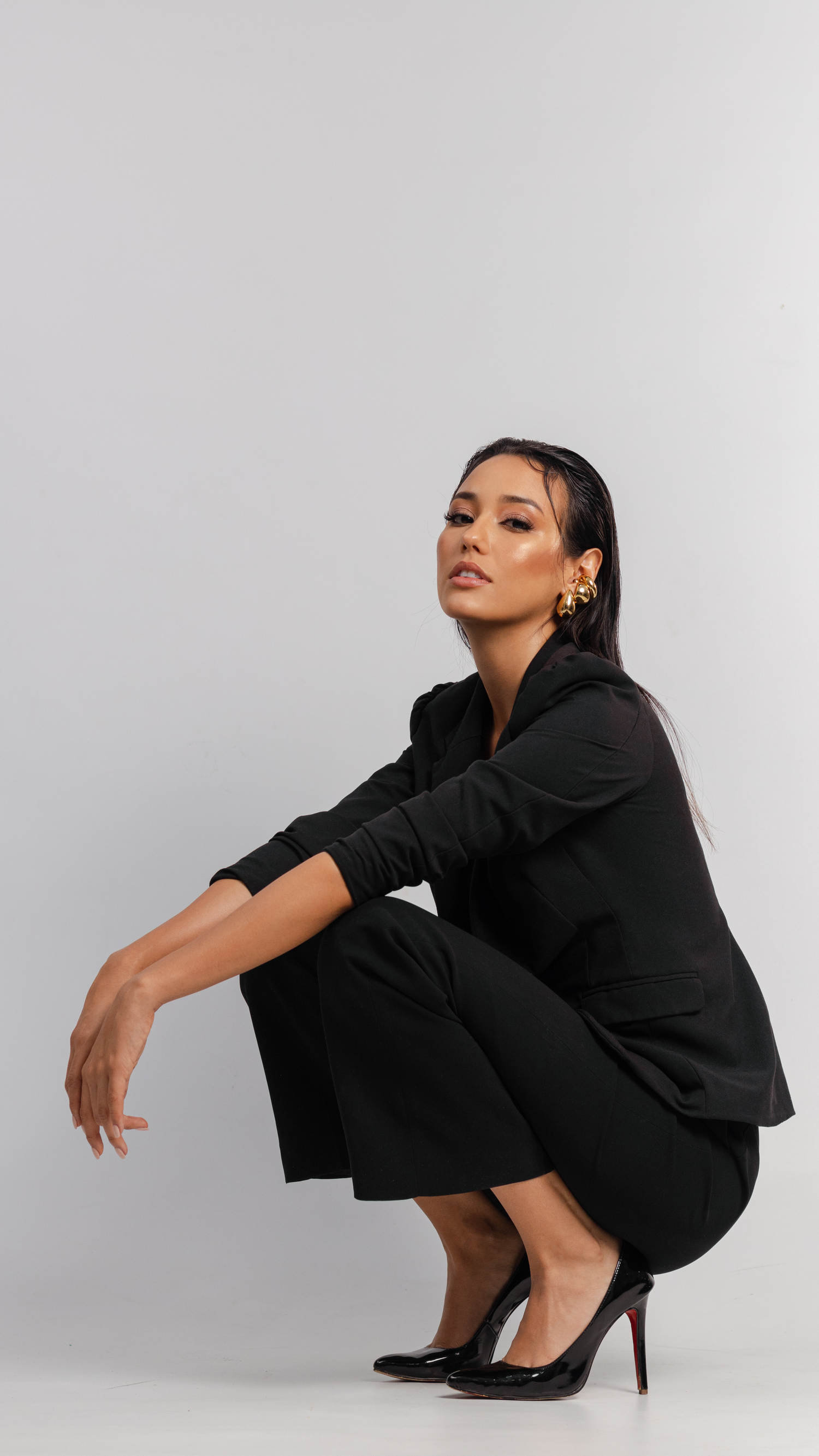 Model in black pantsuit crouches in elegant pose wearing red-bottom stilettos against grey background.