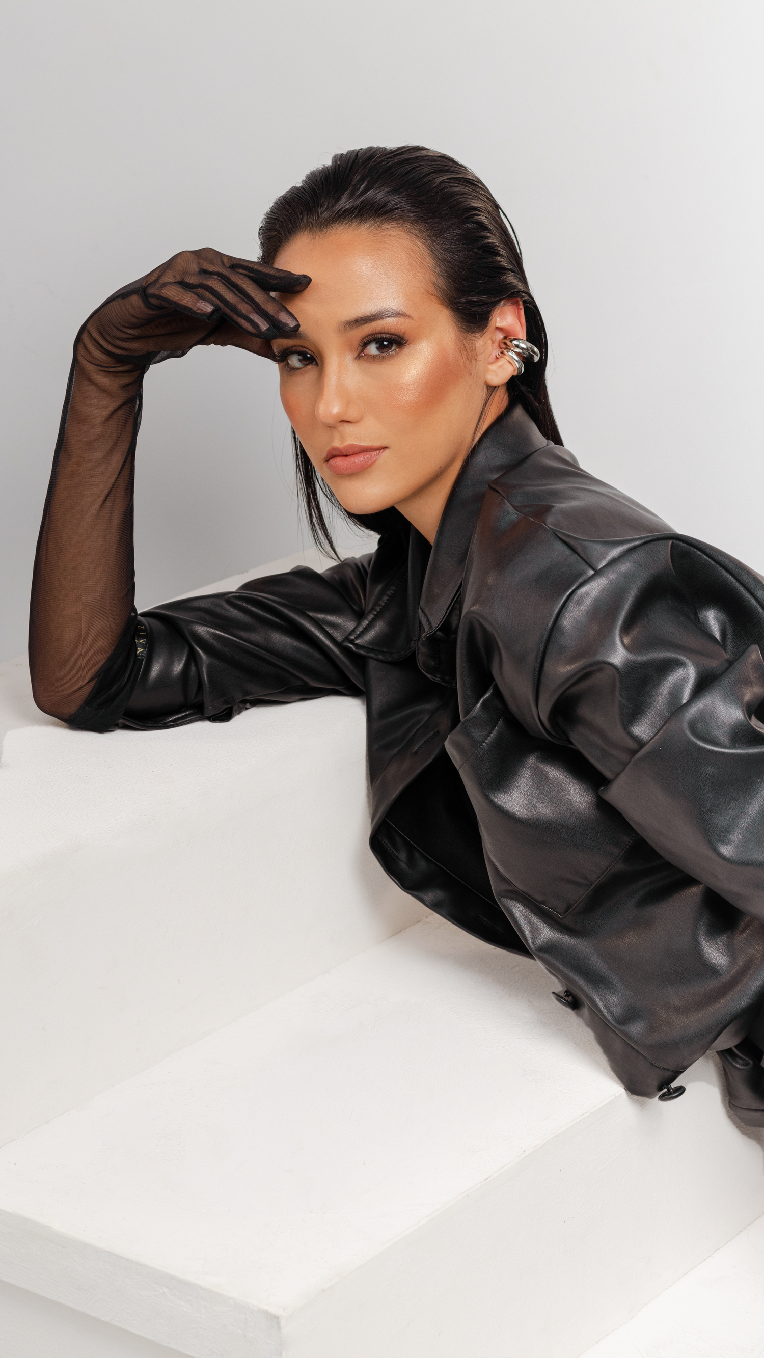 Fashion model in black leather jacket and sheer gloves poses against white background with dramatic lighting.