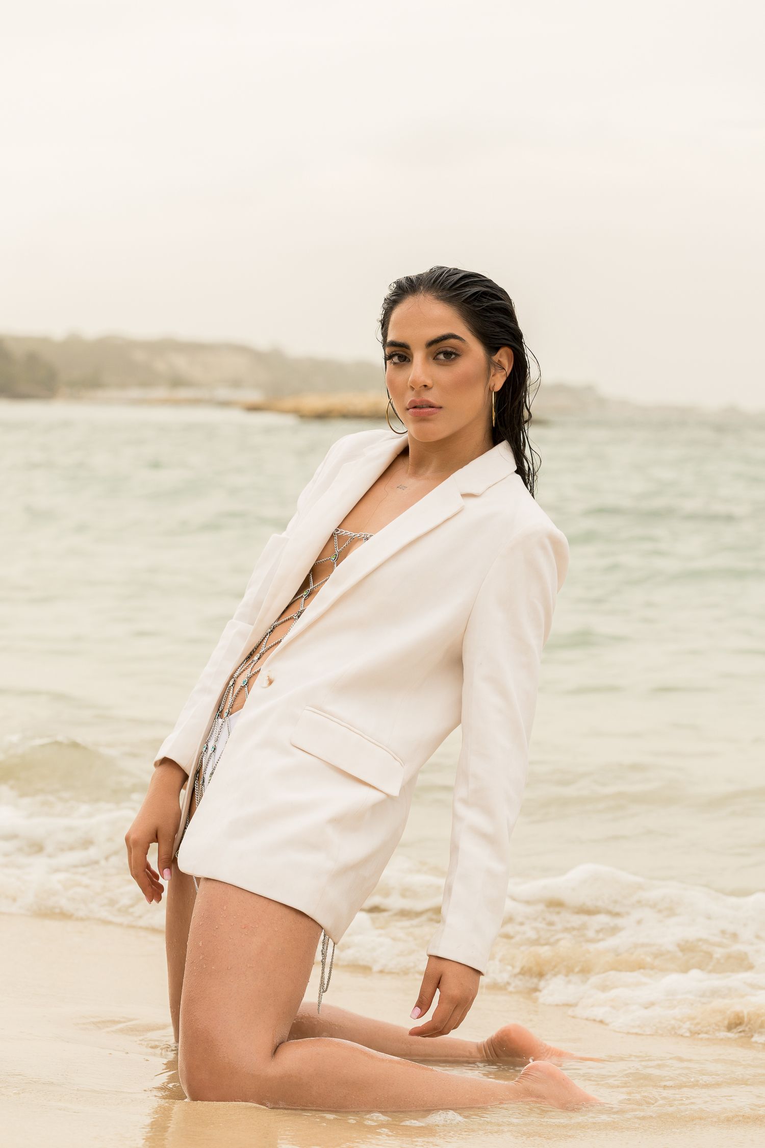 Model in white blazer poses kneeling on sandy beach with overcast sky.