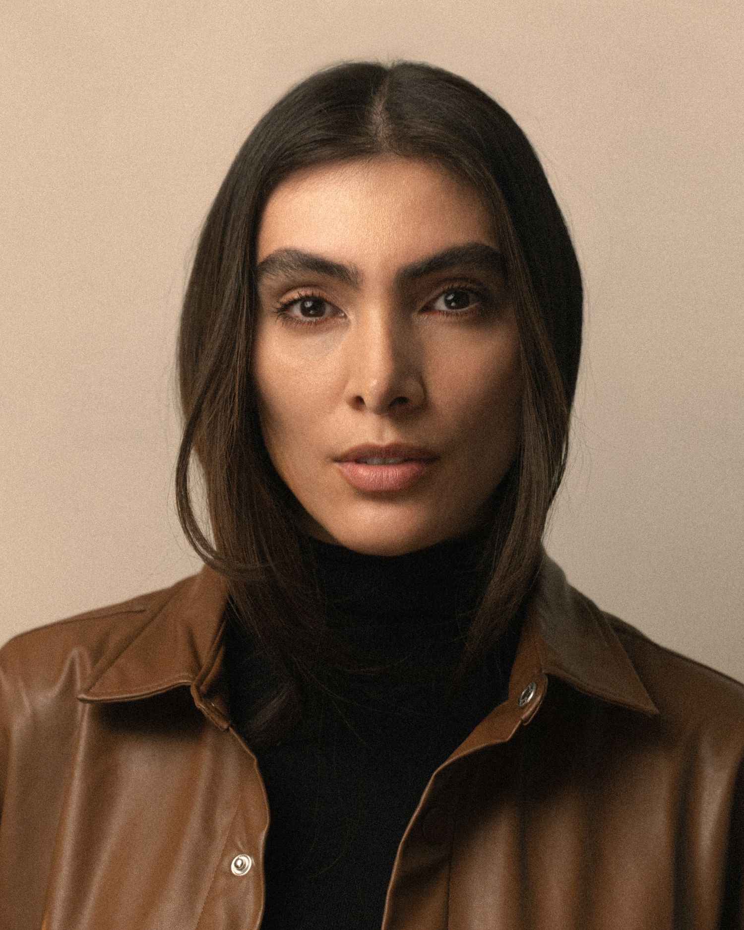 Portrait of someone wearing a black turtleneck and brown leather jacket against a neutral beige background.