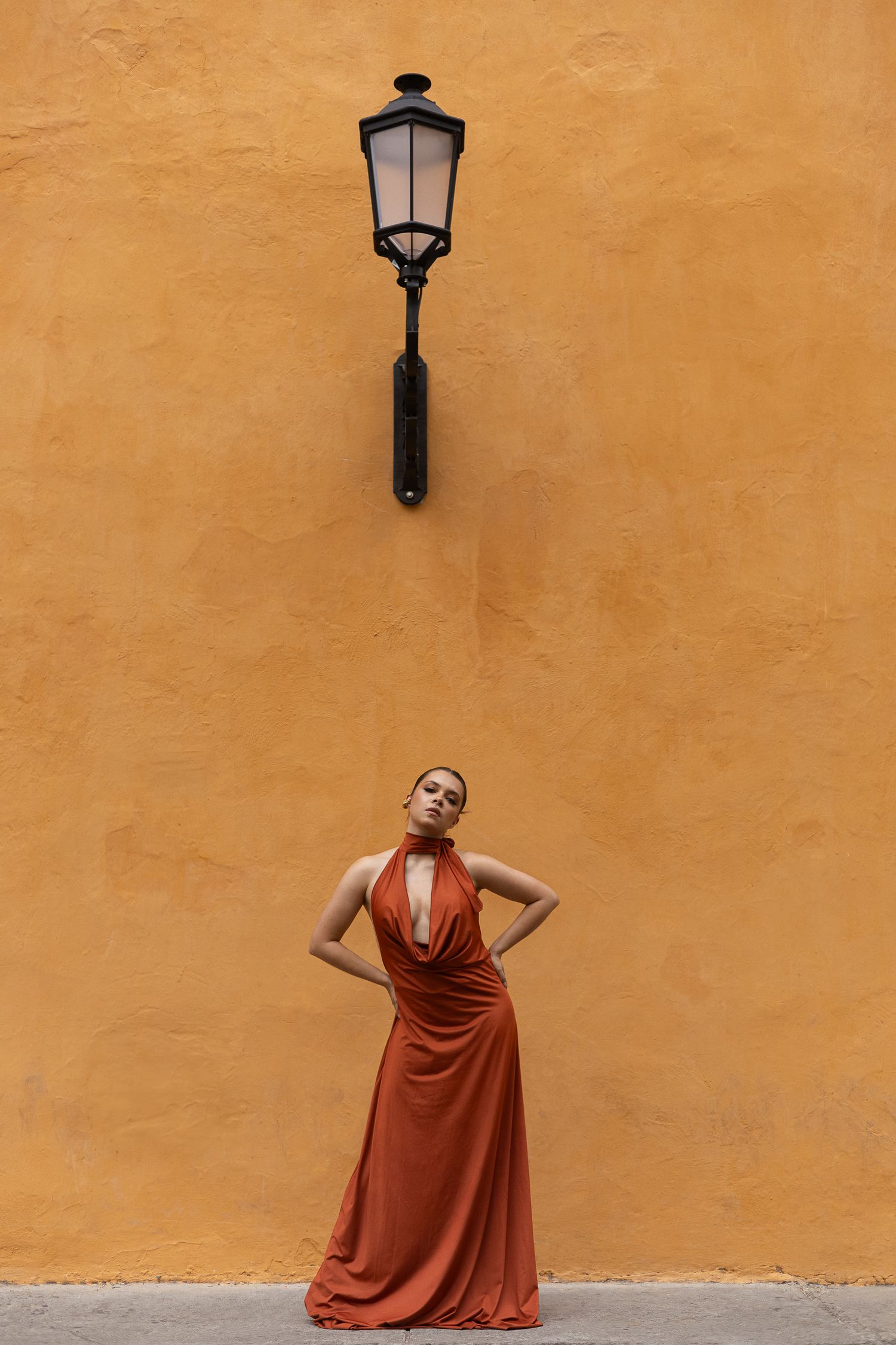 Figure in flowing rust-colored dress poses beneath vintage wall lamp against orange wall.