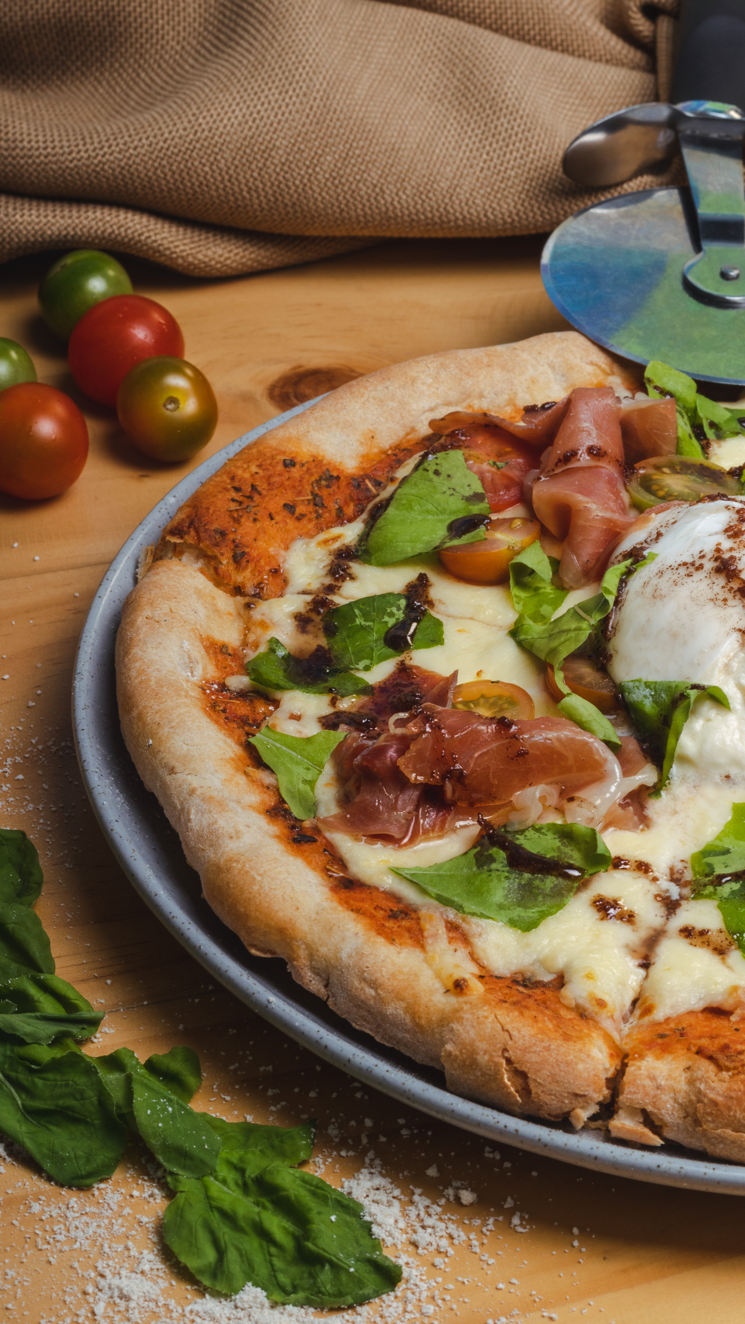 Gourmet pizza topped with prosciutto, burrata, cherry tomatoes and arugula on dark plate.