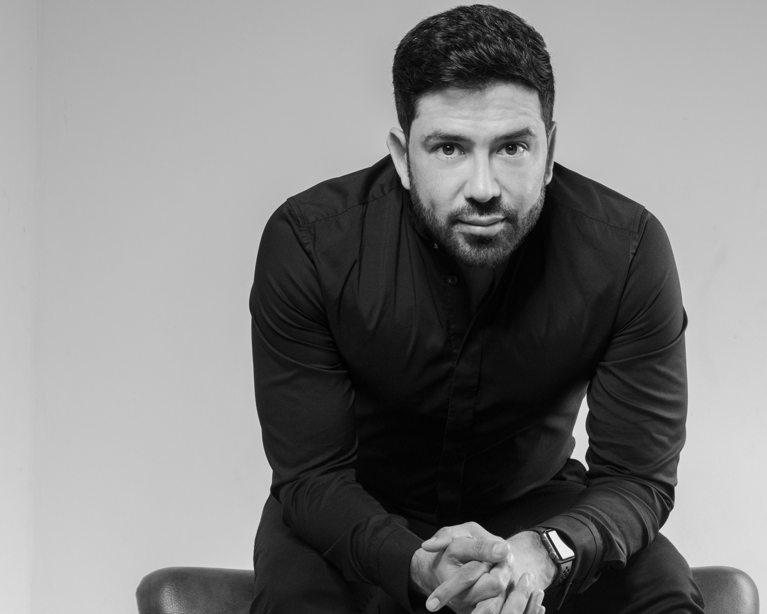 Professional studio portrait of a man in black sweater sitting against grey background.