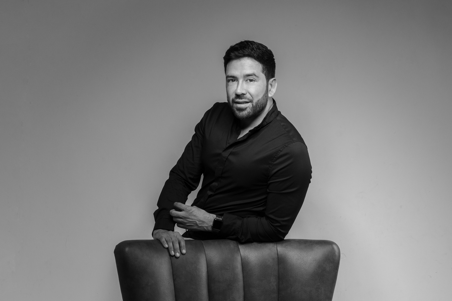 Professional black and white portrait of person in dark shirt posed on leather chair.