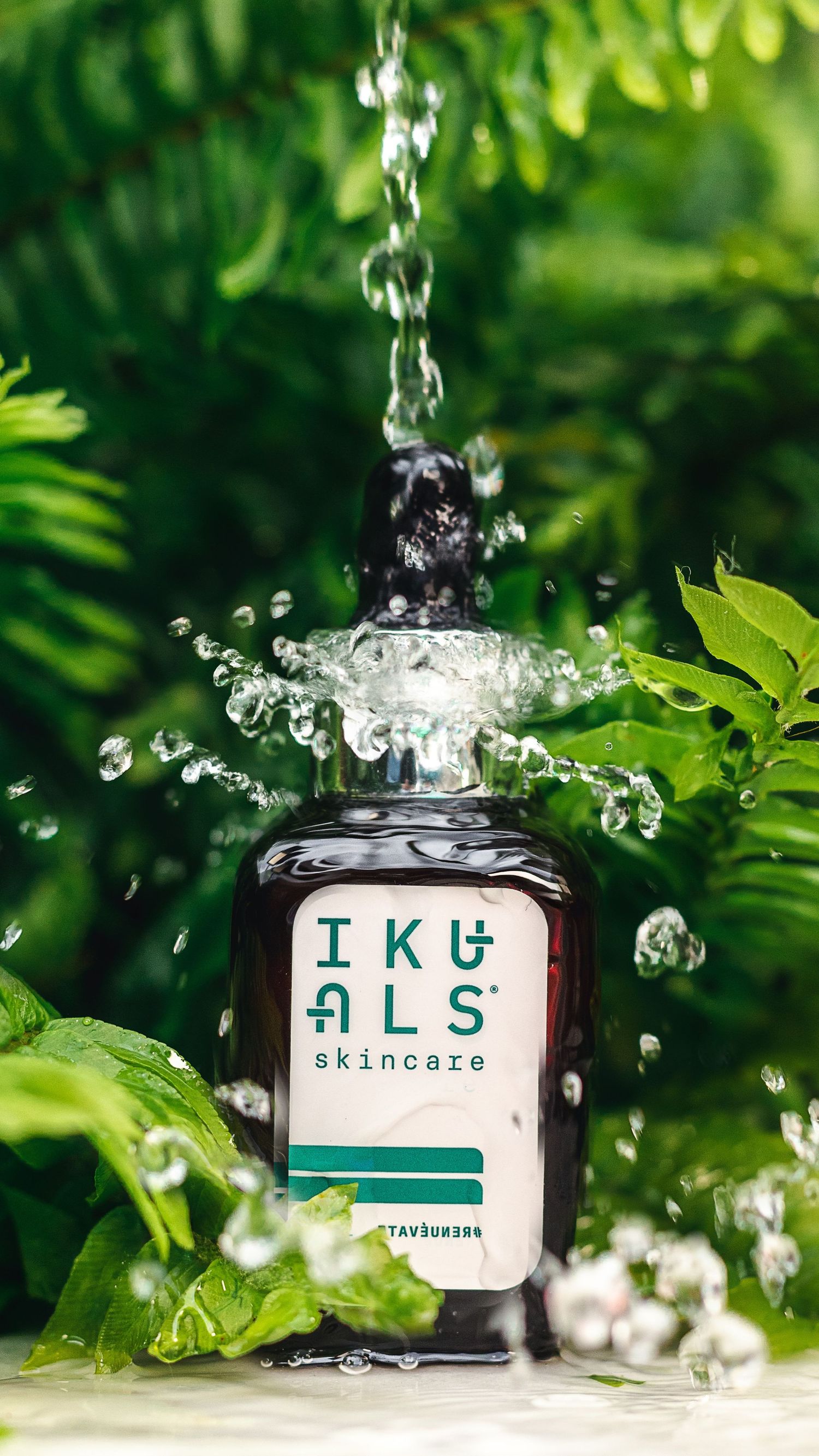 Water splashing dramatically over a dark glass bottle of skincare product among green foliage.