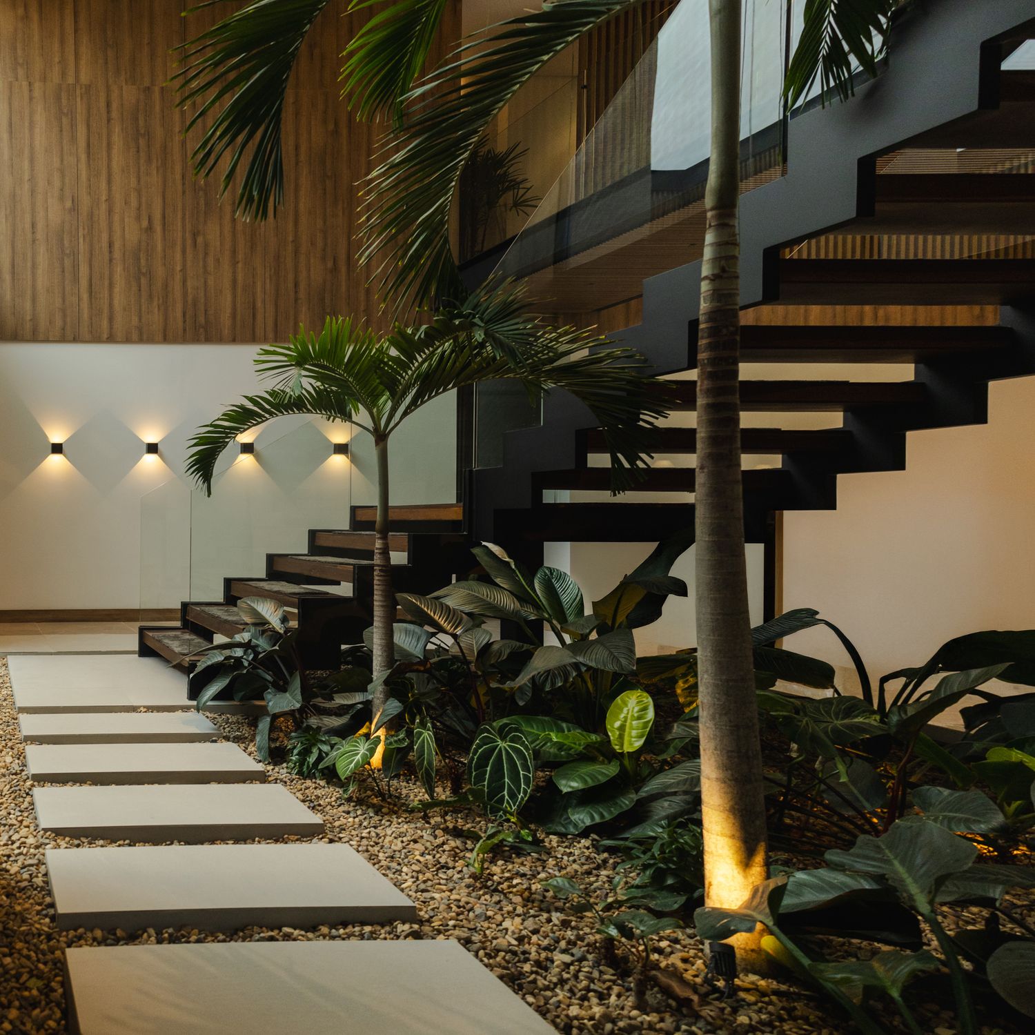 Modern floating staircase with lush tropical plants, stone pathway, and warm wall lighting in a luxury interior.